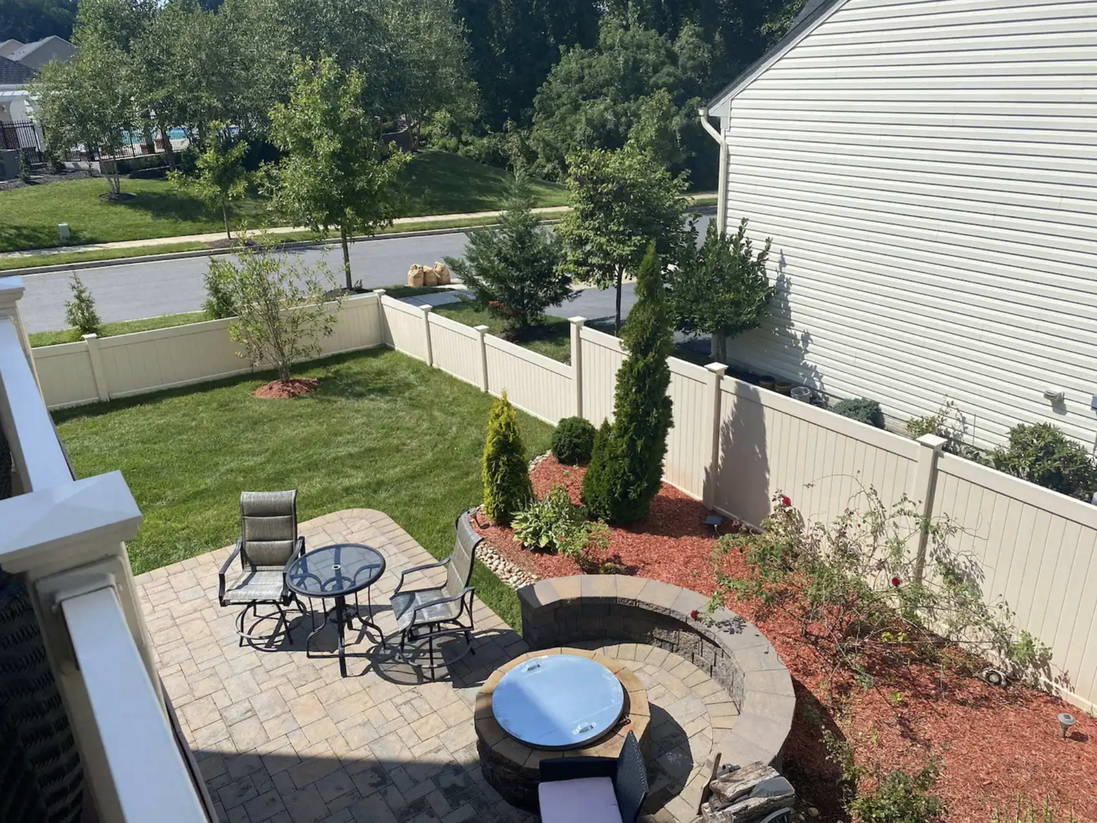 The image shows a landscaped backyard featuring a stone patio, a fire pit, seating area with metal chairs, and a green lawn bordered by a white fence.
