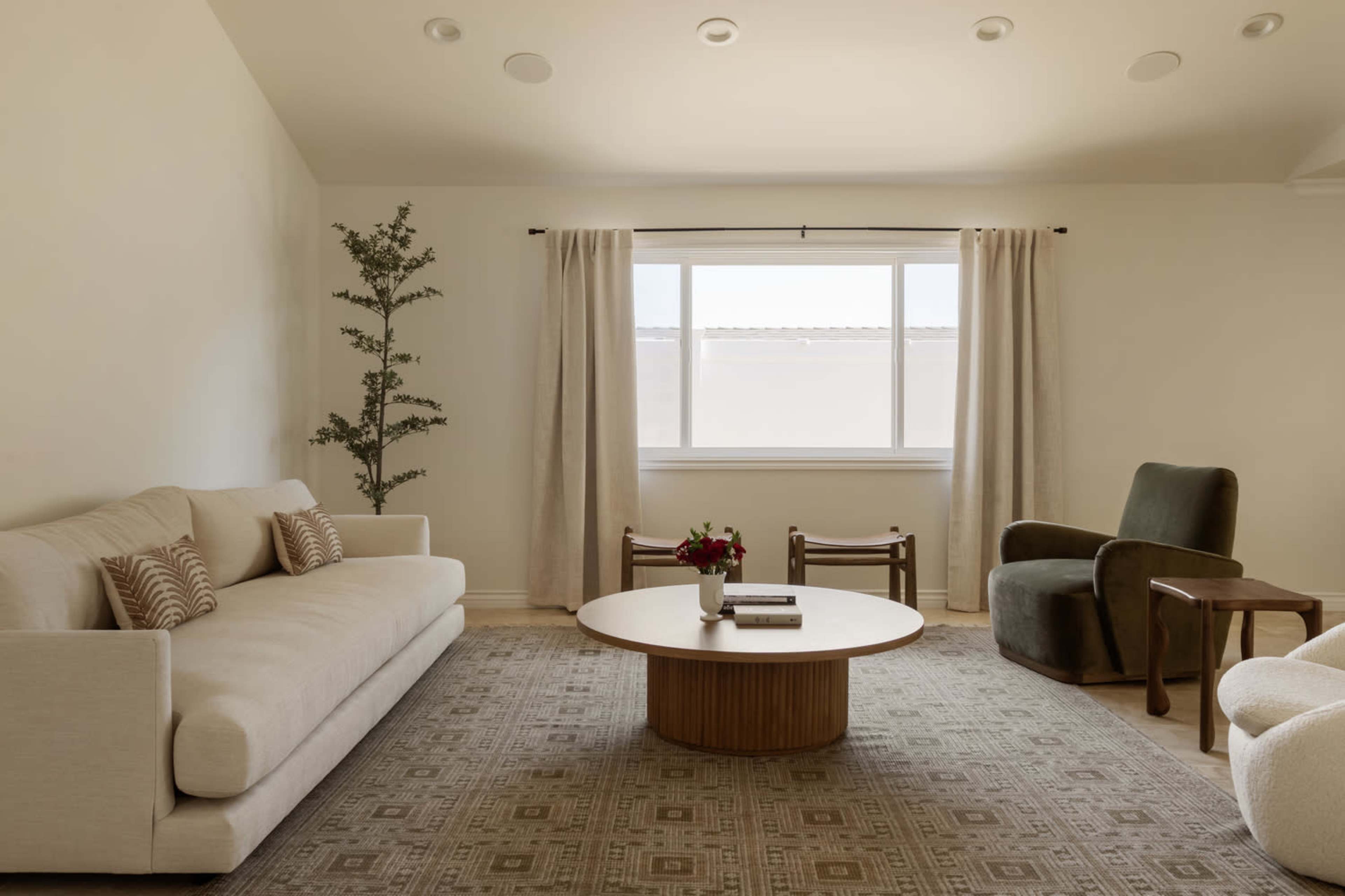 A minimalist living room features a light-colored sofa, a round coffee table, two armchairs, and a window with sheer curtains allowing natural light to enter.