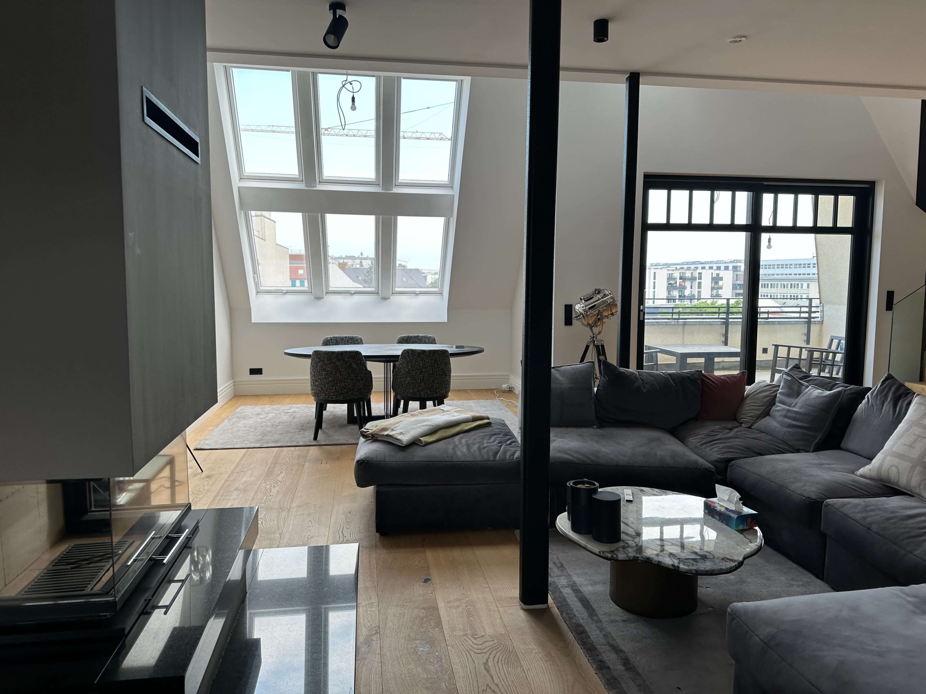 The image shows a modern, open living space featuring a sectional sofa, a glass coffee table, and a dining area with a table and chairs, illuminated by natural light from large windows.