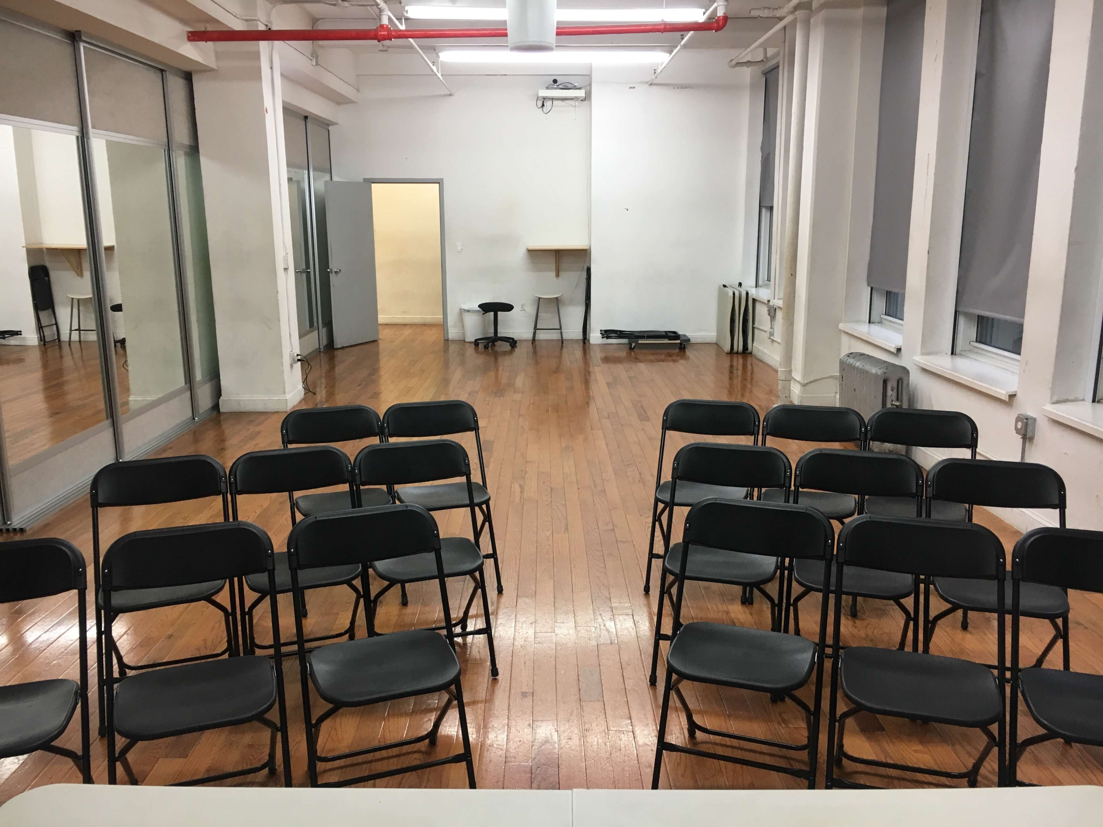 The image shows a room with wooden flooring, featuring rows of black chairs arranged facing a table at the front, with additional empty space and a doorway in the background.