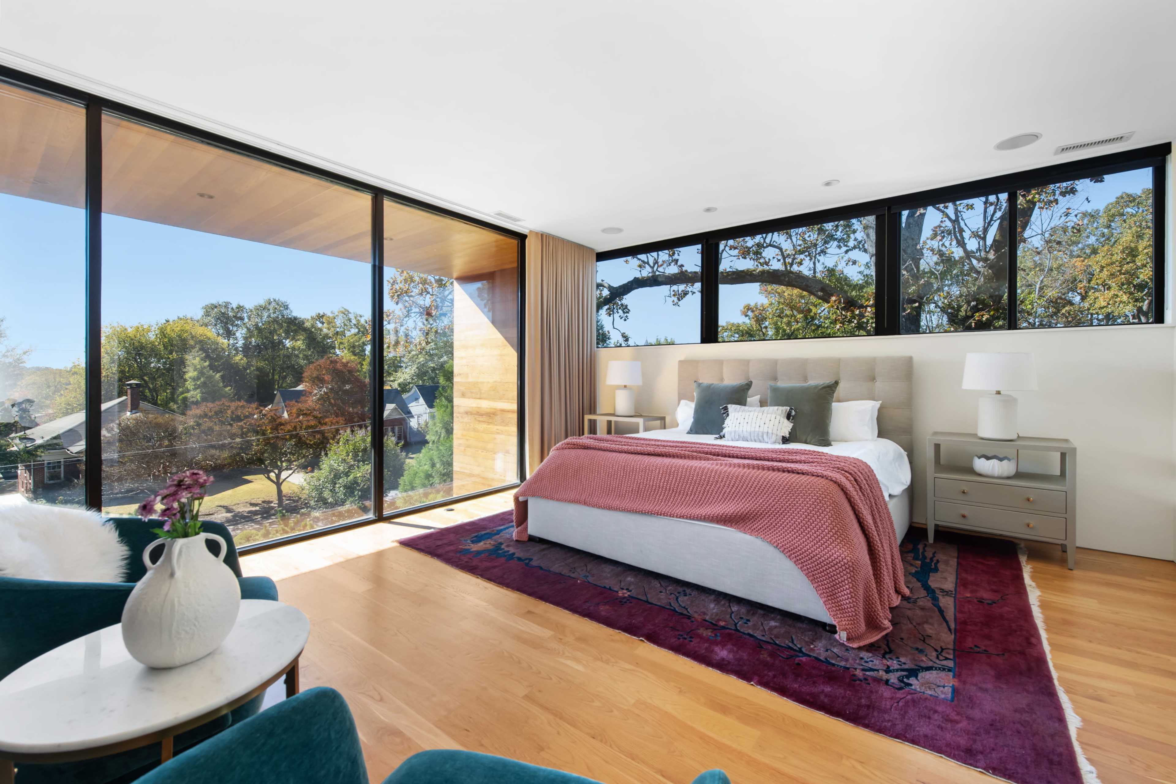 The image shows a modern bedroom with large windows allowing natural light and offering a view of the outdoors, featuring a bed with a red blanket and a rug on wooden flooring.