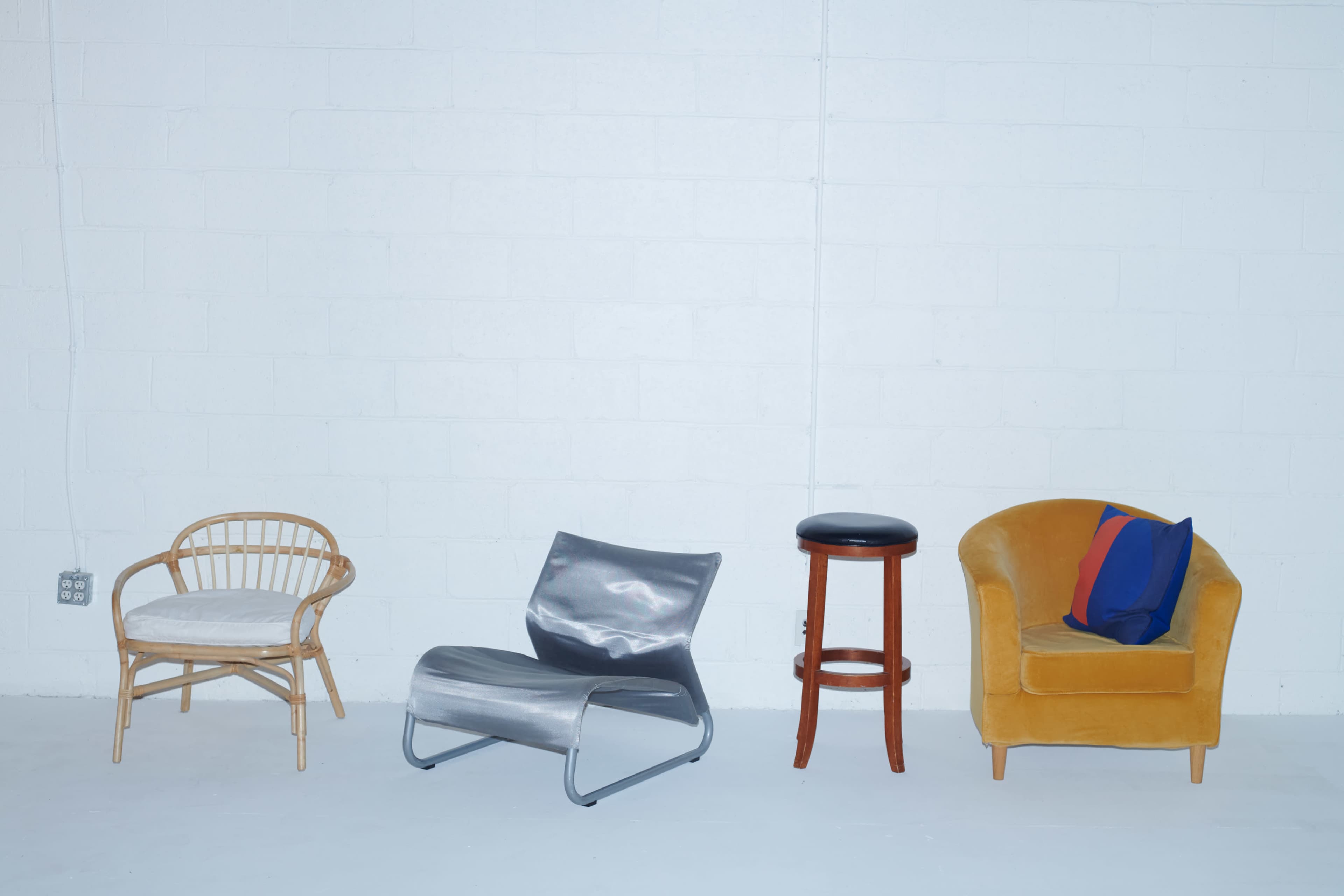 Four distinct chairs arranged against a plain white wall, including a rattan chair, a metallic lounge chair, a wooden stool, and a yellow upholstered armchair with a blue and red pillow.