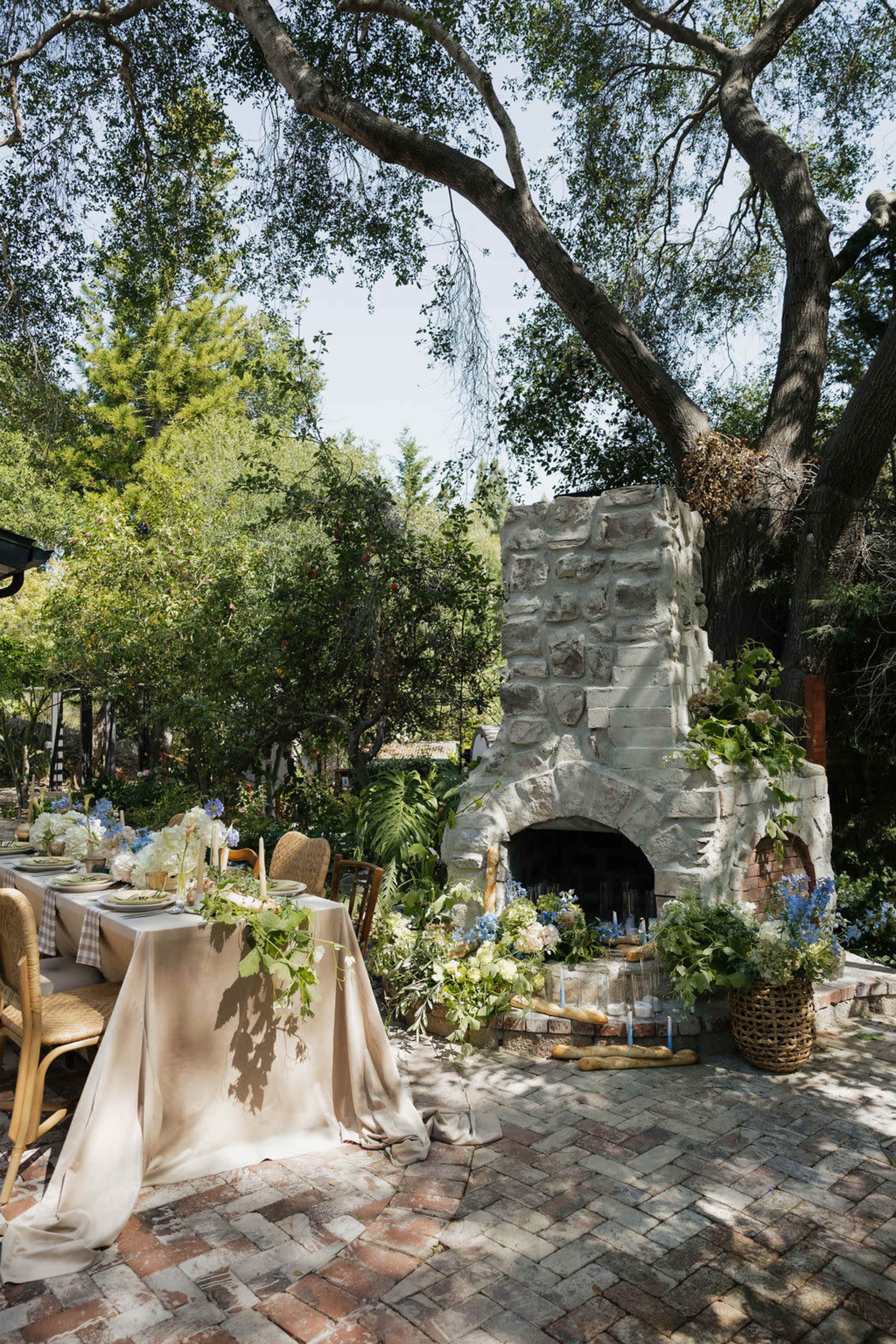 A rustic outdoor dining area features a stone fireplace surrounded by greenery and a table set with natural elements.