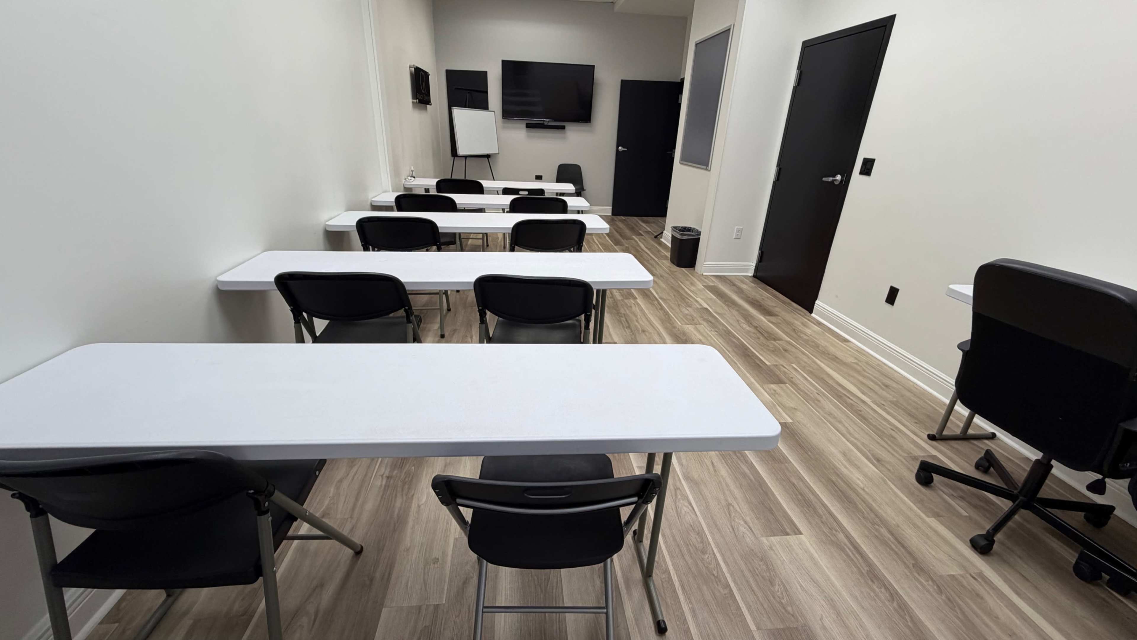 The image shows a small classroom with several white folding tables and black chairs arranged in rows, featuring a large screen mounted on the wall.
