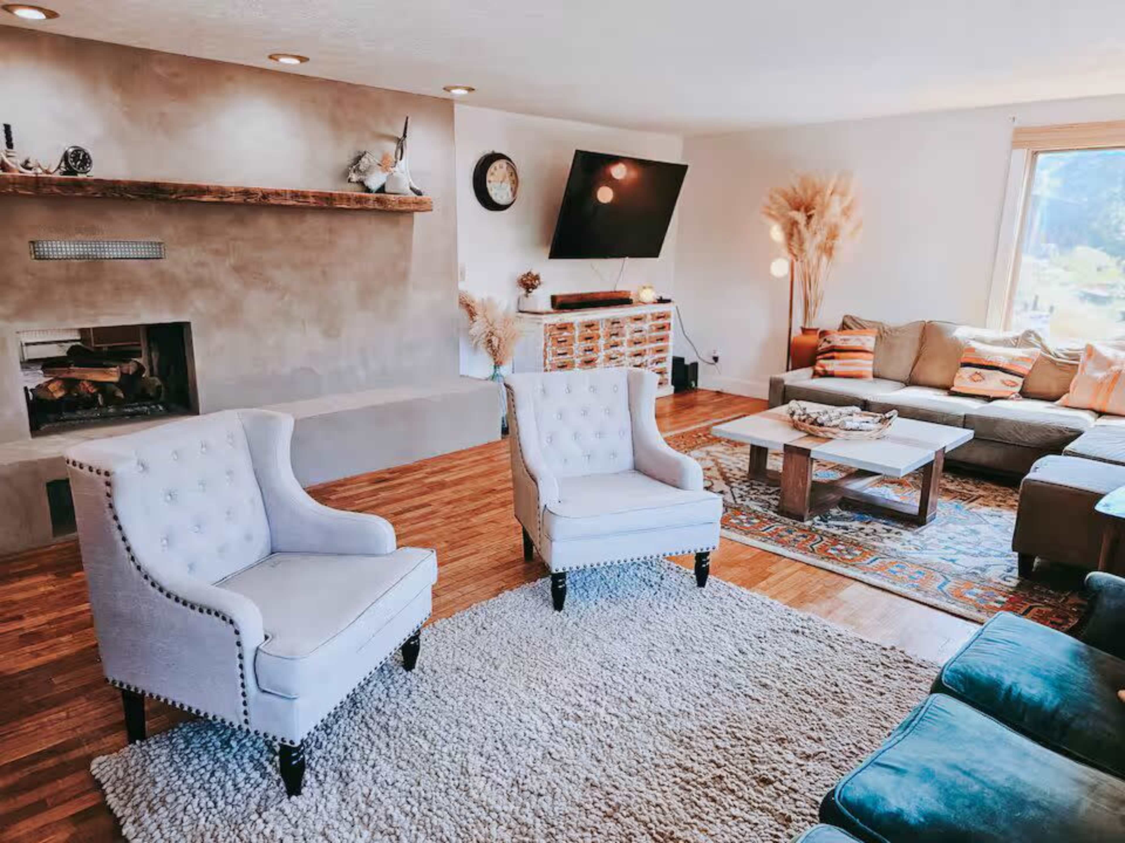 The image shows a modern living room featuring two upholstered chairs, a coffee table, a sofa, and a wall-mounted television, with warm wooden floors and natural light coming through a large window.