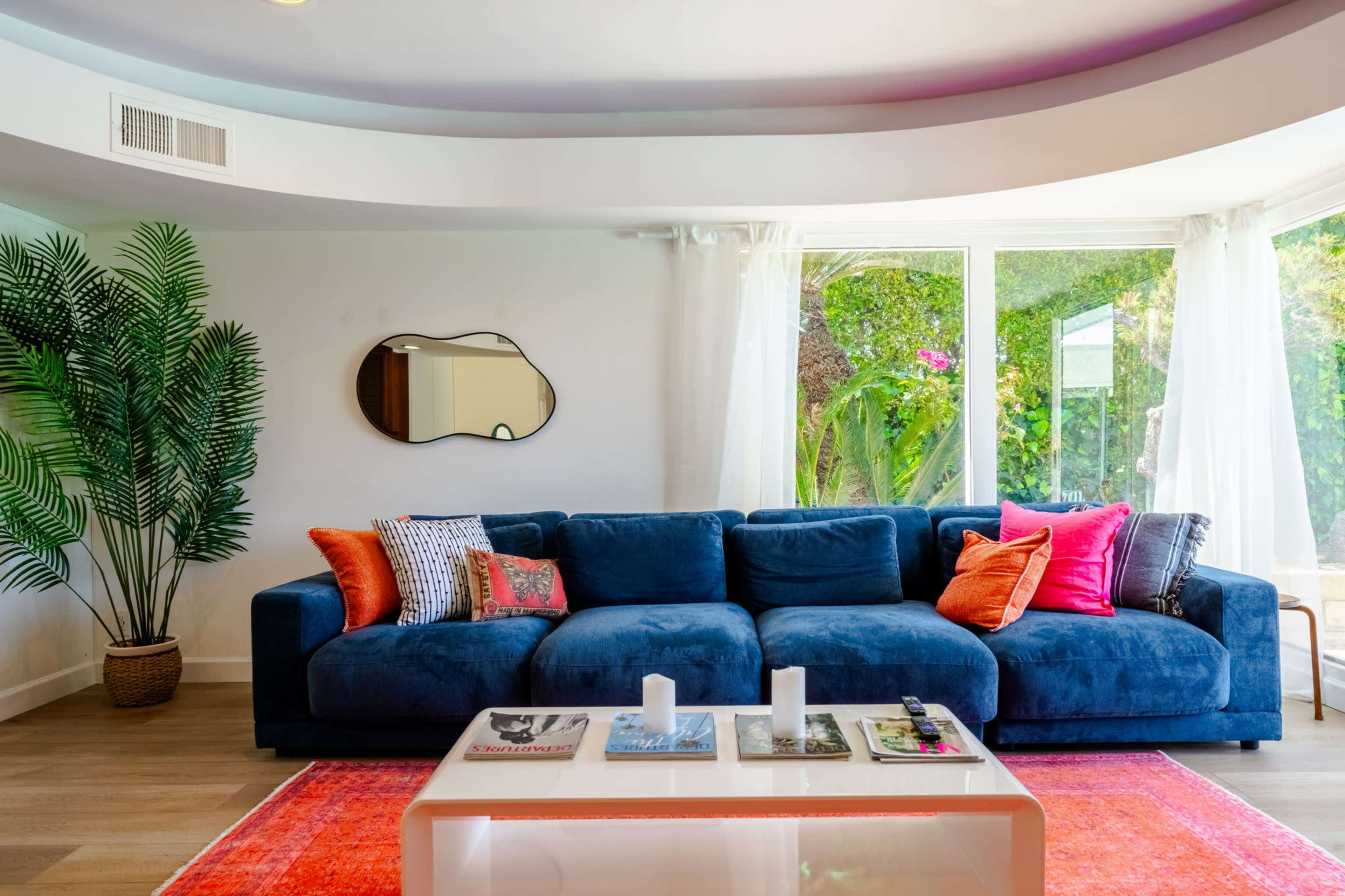 A spacious living room features a large blue sectional sofa adorned with colorful cushions, a glass coffee table with magazines, and a plant positioned next to a window.