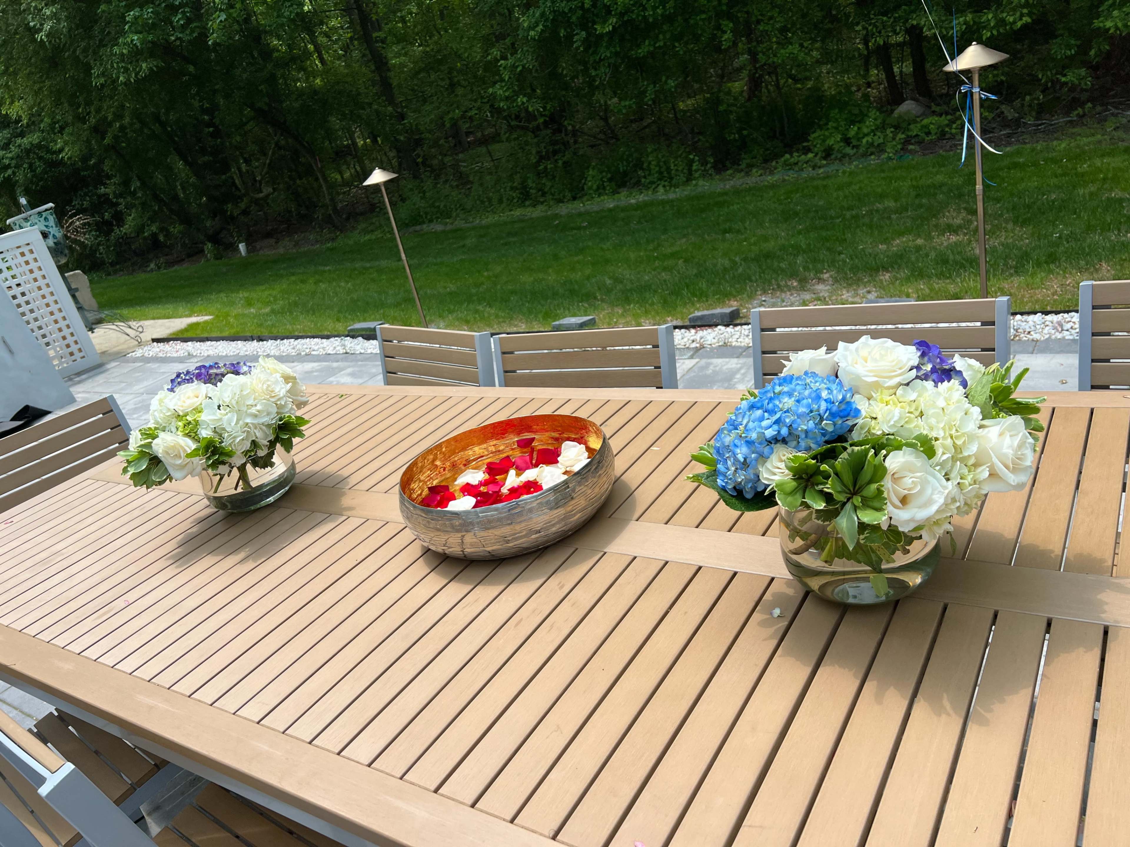 The outdoor dining table is set with two flower arrangements and a decorative bowl filled with petals.