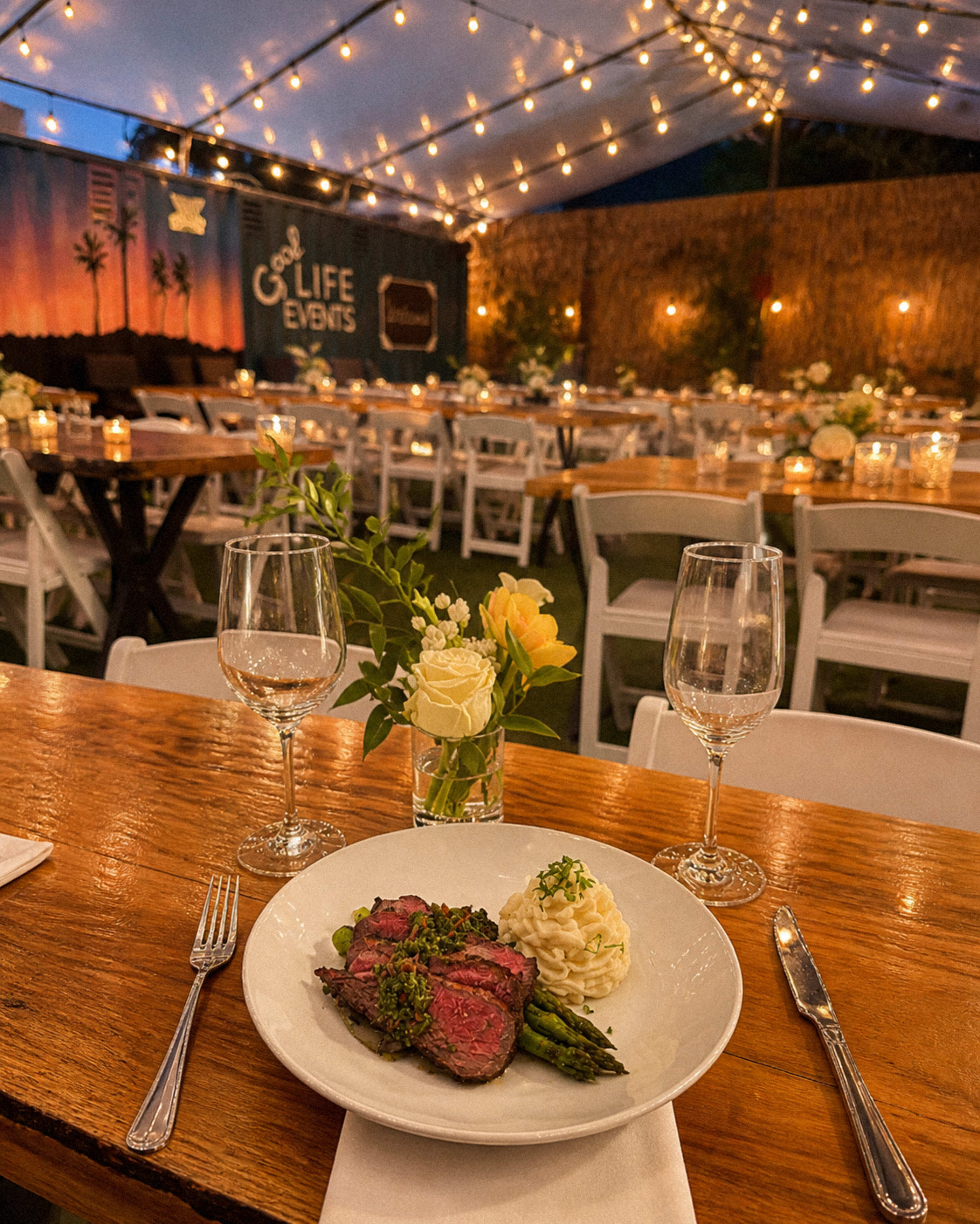 The image shows a beautifully set dining table at an outdoor event, featuring a plate of sliced steak with asparagus and mashed potatoes, alongside wine glasses and floral centerpieces under string lights.