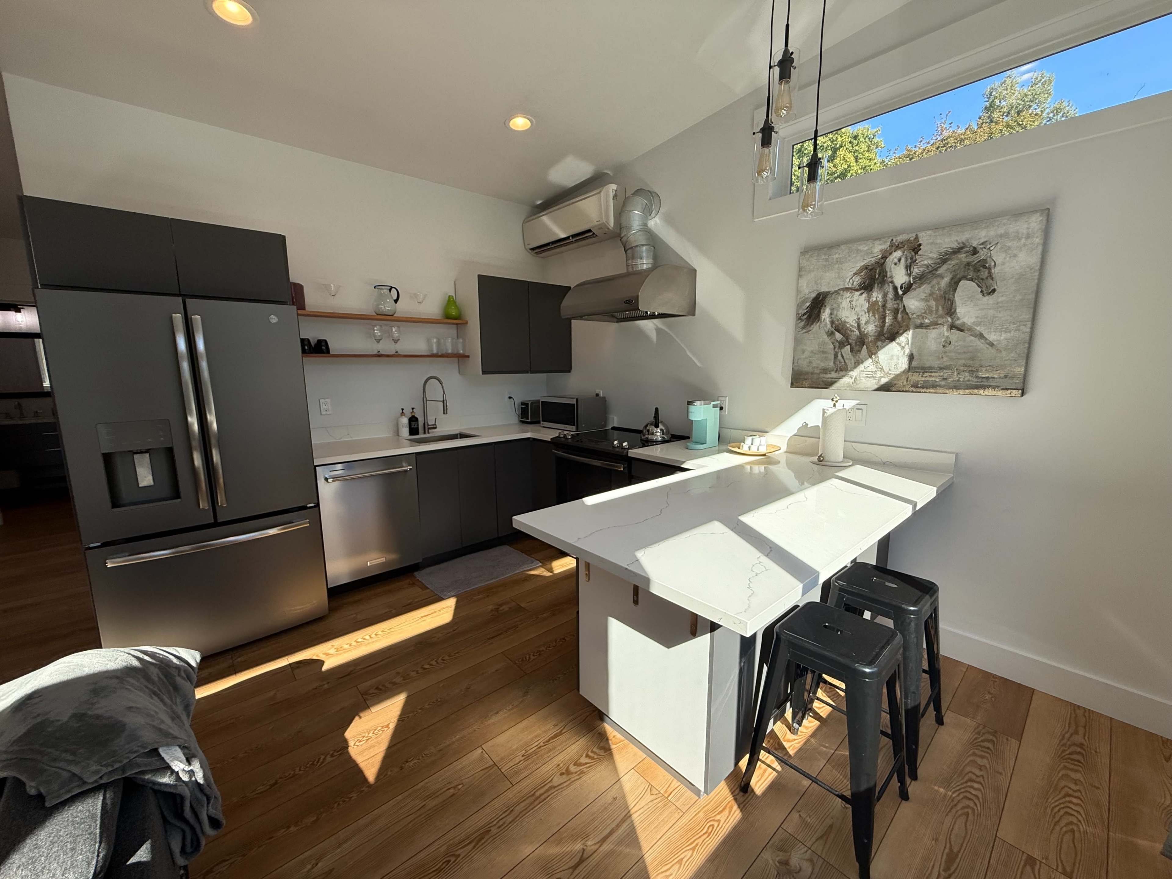 A modern kitchen featuring stainless steel appliances, a white marble island with black stools, and large windows providing natural light.