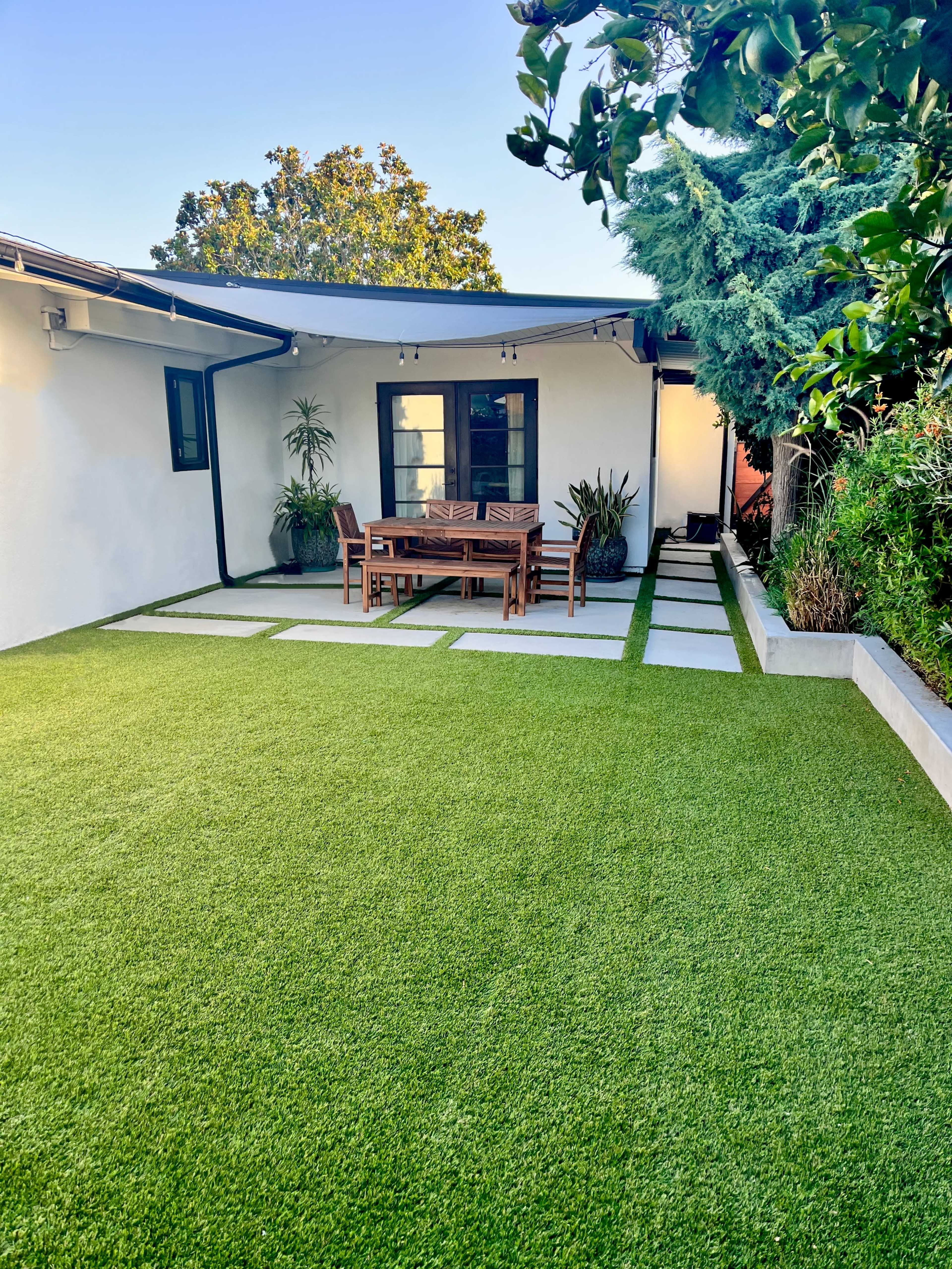A backyard patio featuring a wooden dining table and chairs on a well-maintained artificial lawn, adjacent to a house with a modern exterior.