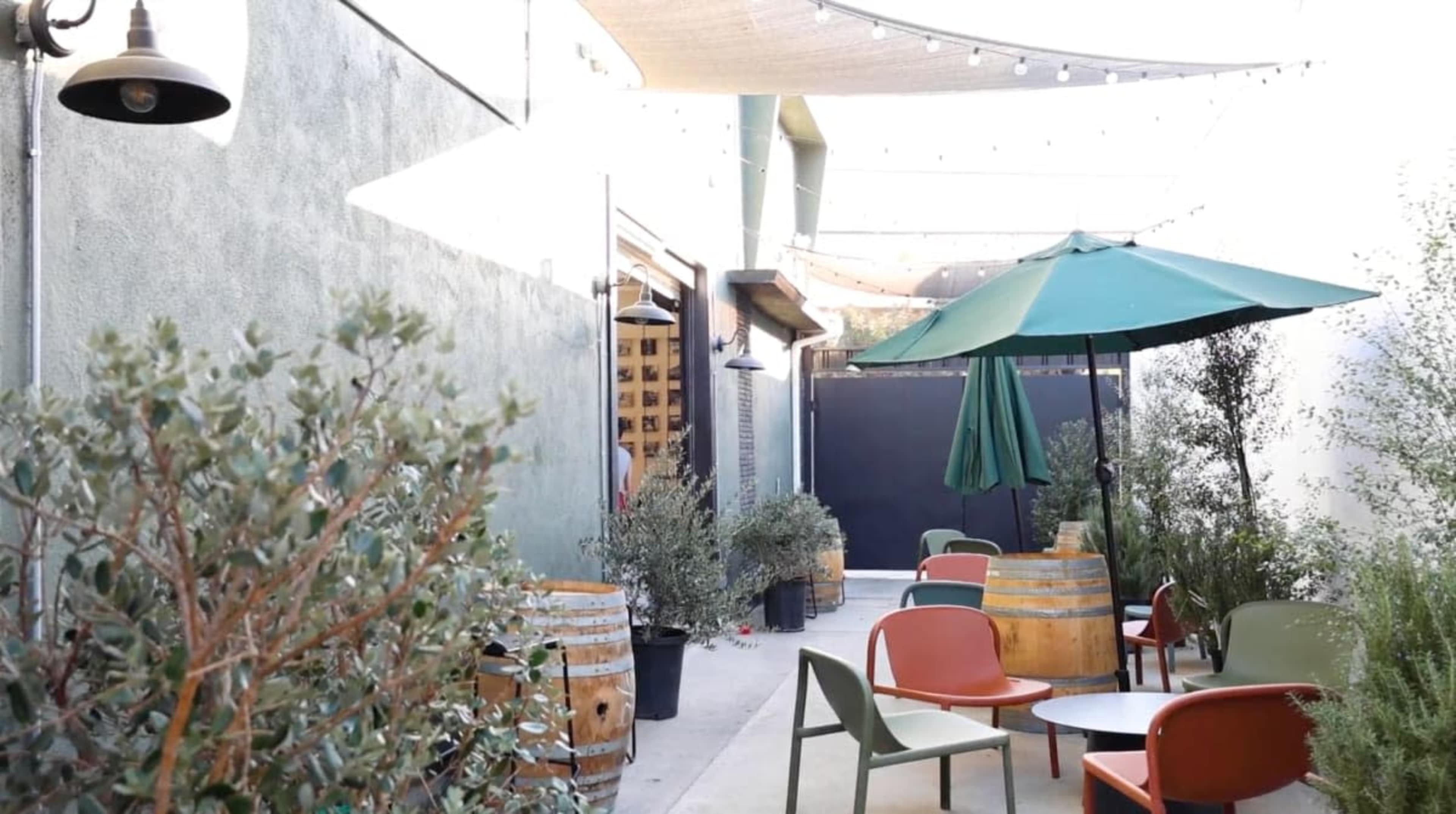 The image shows a narrow outdoor seating area with green umbrellas, colorful chairs, and potted plants, illuminated by overhead string lights.