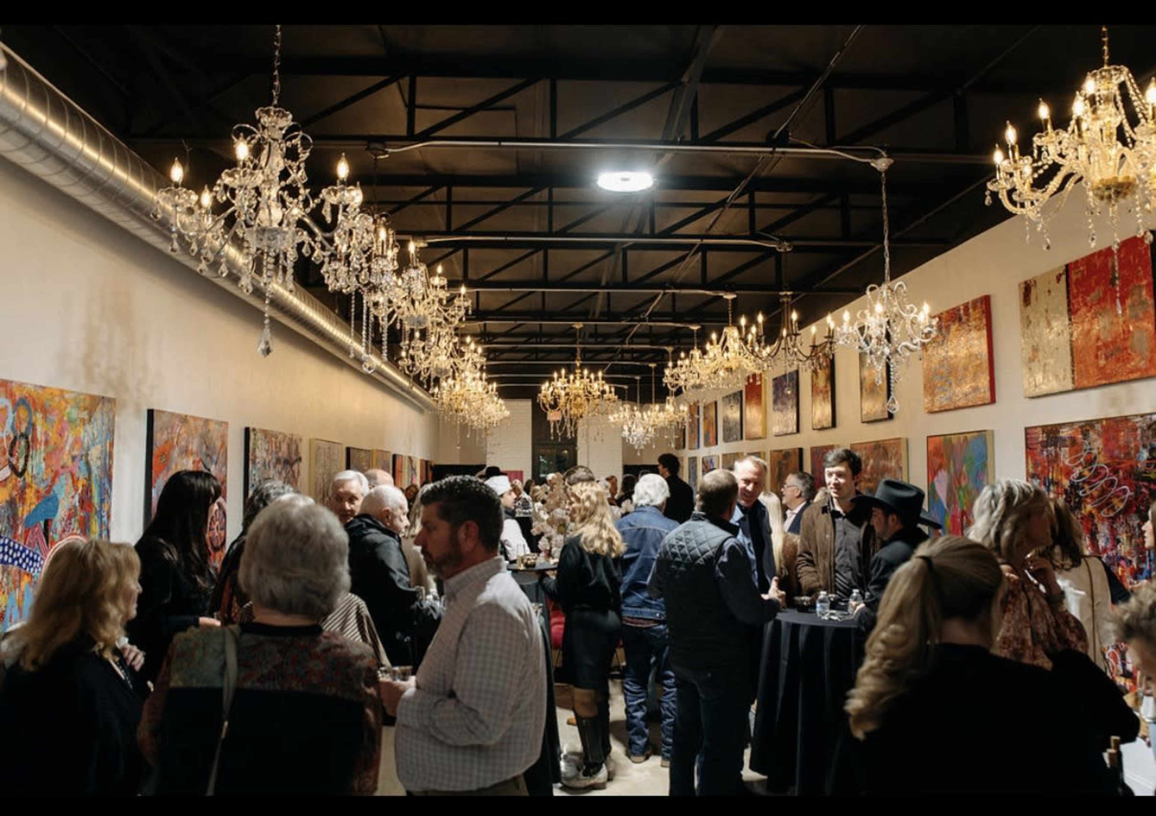 A crowded art gallery features people mingling under ornate chandeliers while colorful artwork adorns the walls.