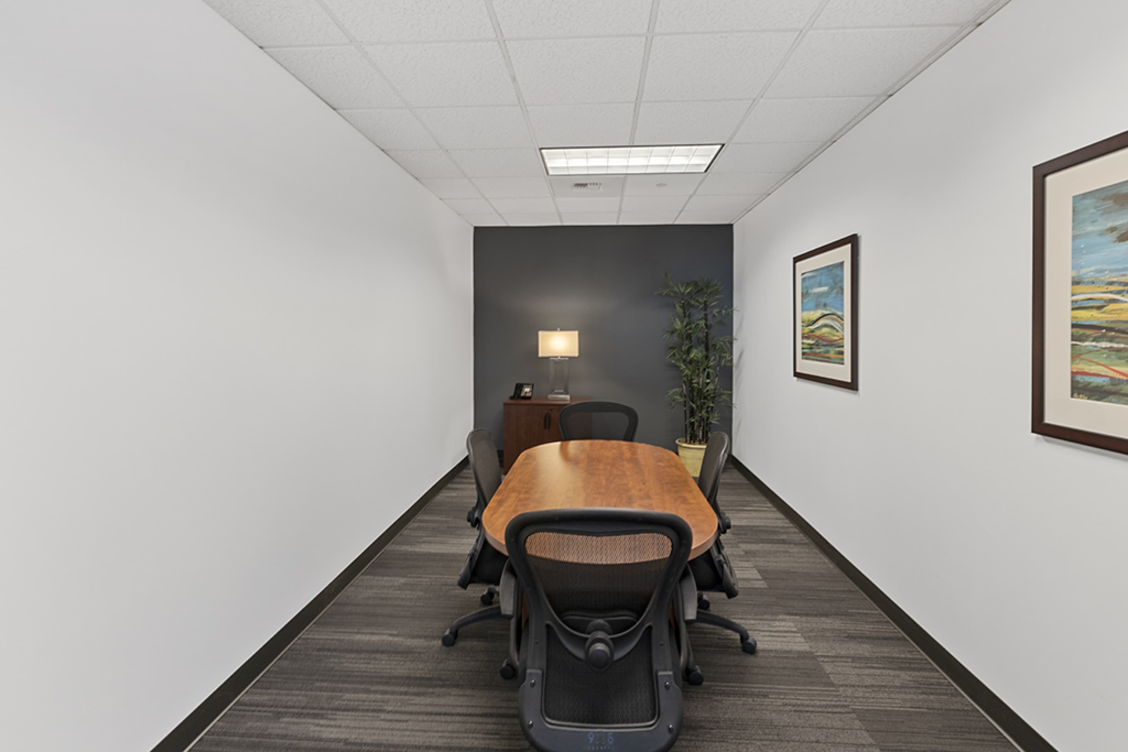 A long conference room with a wooden table, black office chairs, a lamp on a desk, and framed artwork on the walls.