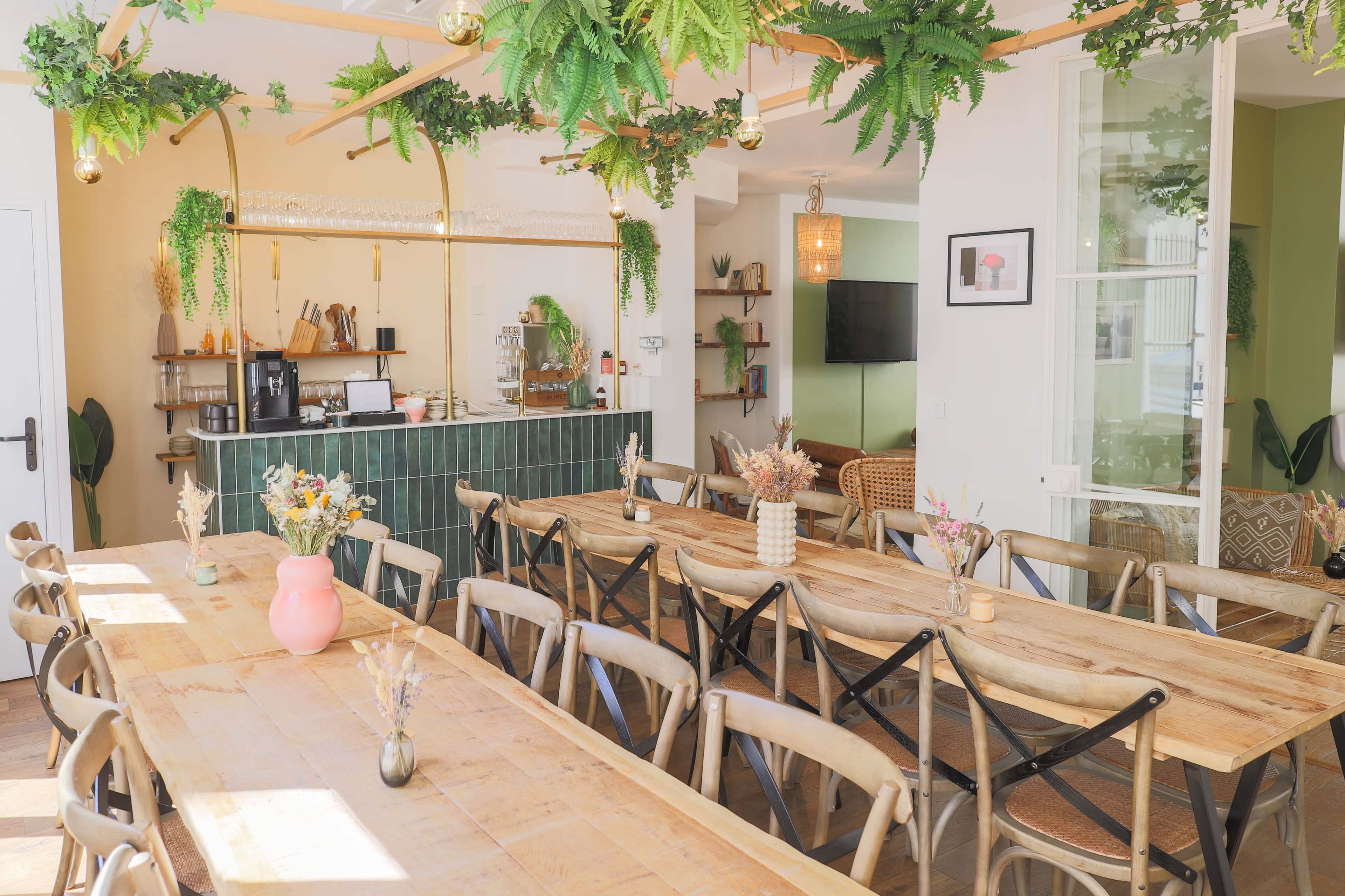 A spacious café features a long wooden table surrounded by chairs, with greenery hanging from the ceiling and a counter in the background.