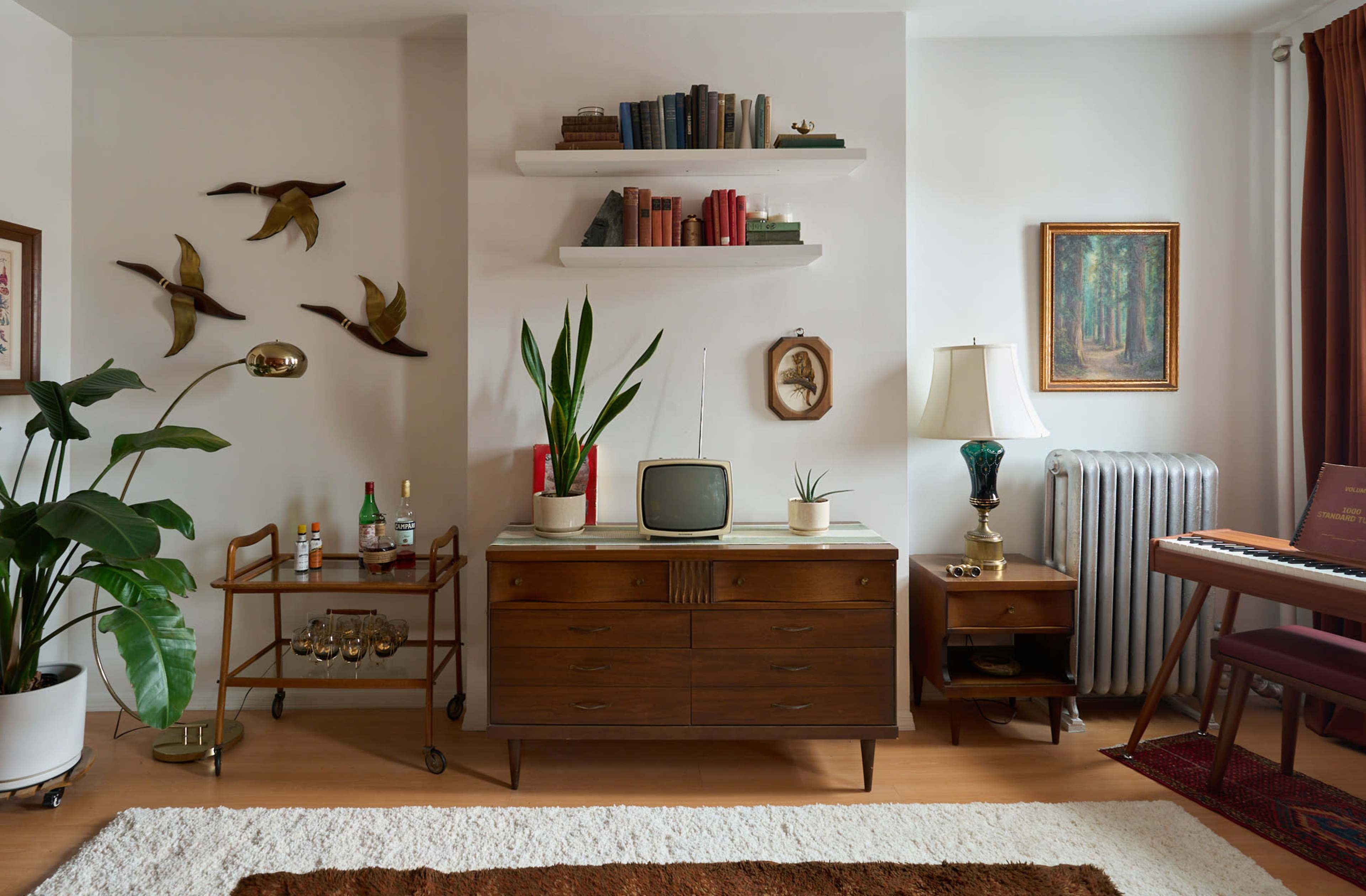 The image depicts a stylish retro living room featuring a wooden sideboard with a vintage television, a bar cart, potted plants, and various books displayed on shelves.
