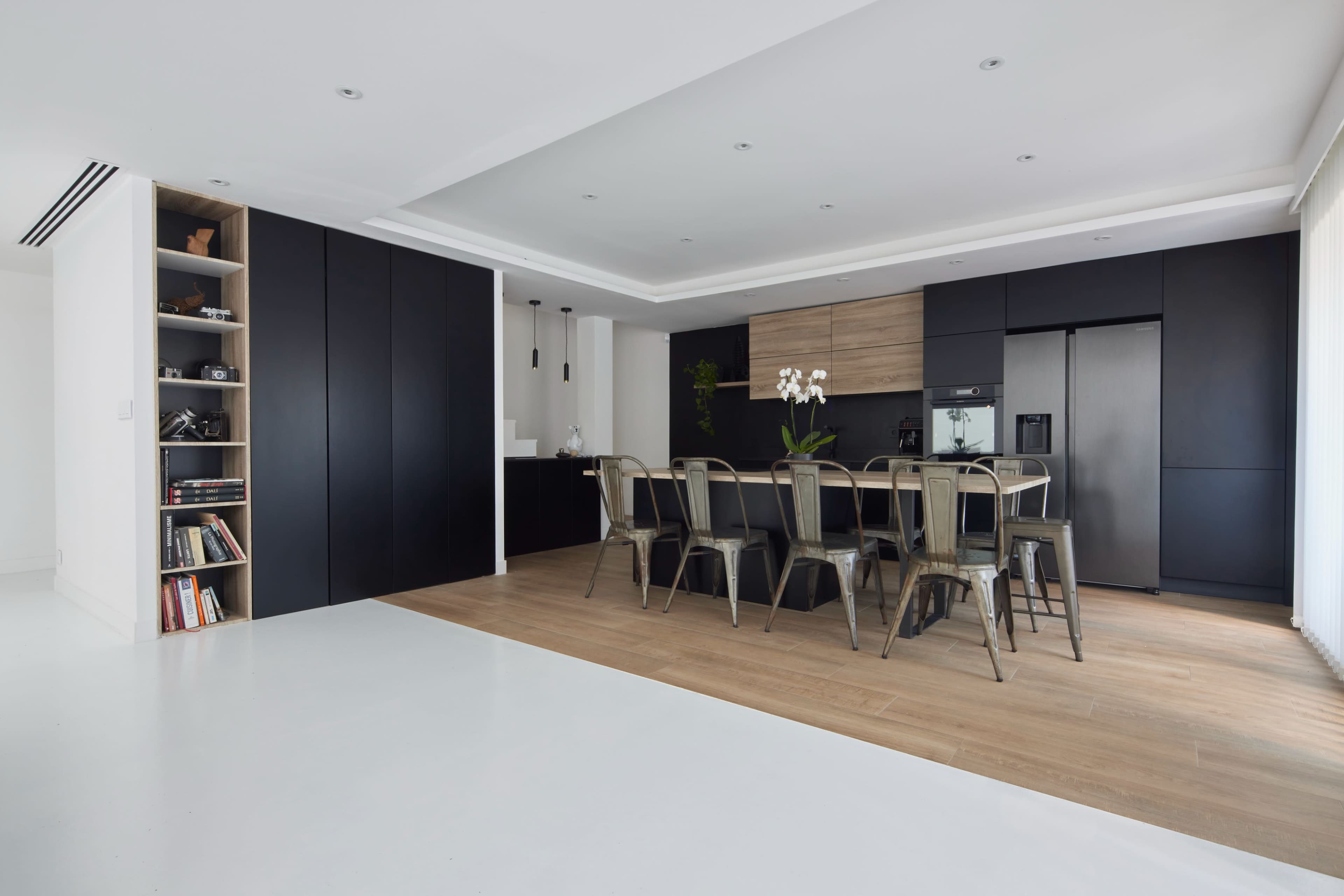 A modern kitchen and dining area features a sleek design with dark cabinetry, a wooden accent wall, and a large table surrounded by metal chairs.