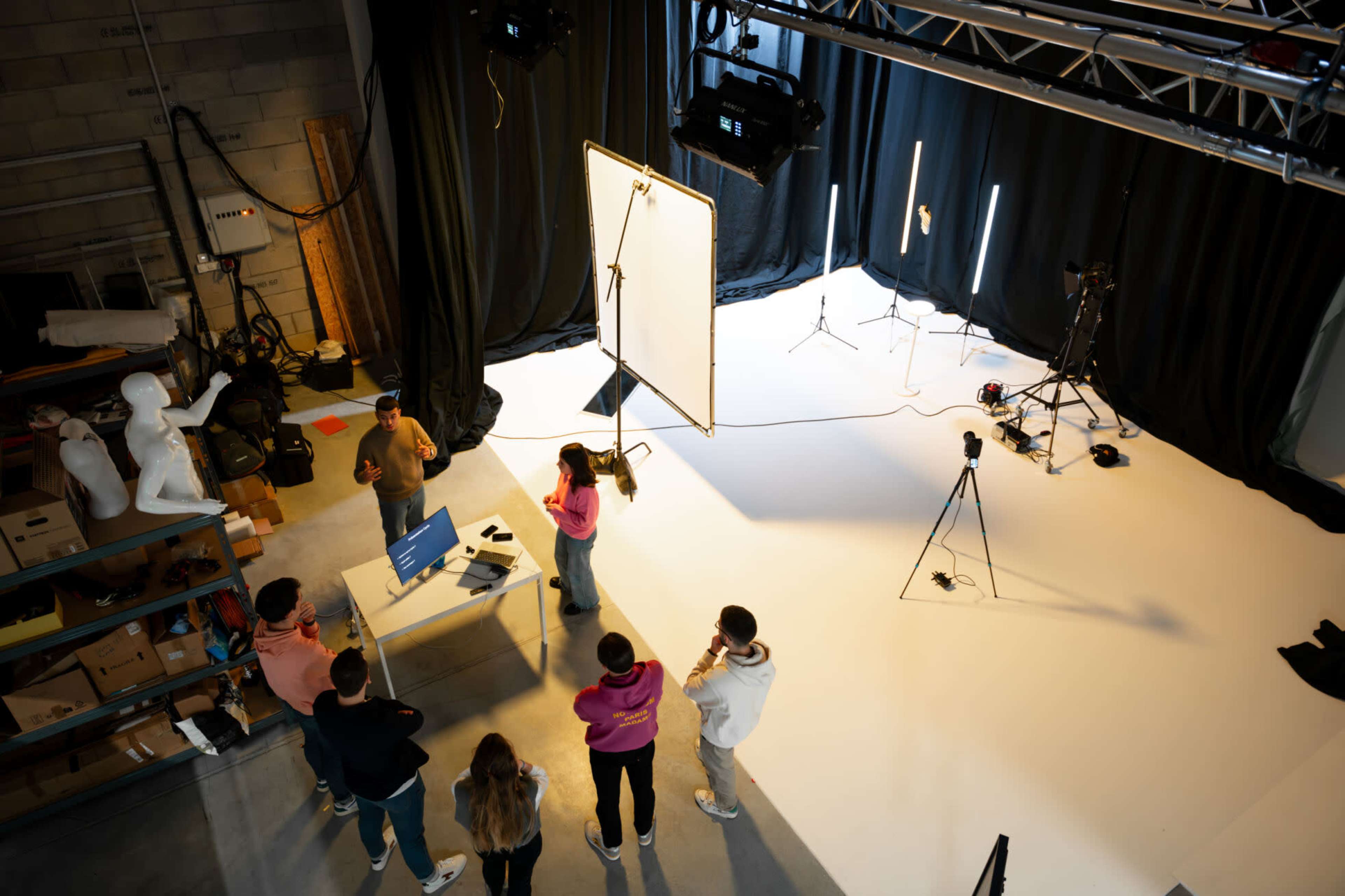 A group of individuals stands around a table with a laptop in a photography studio, while various lighting and camera equipment are positioned around them.