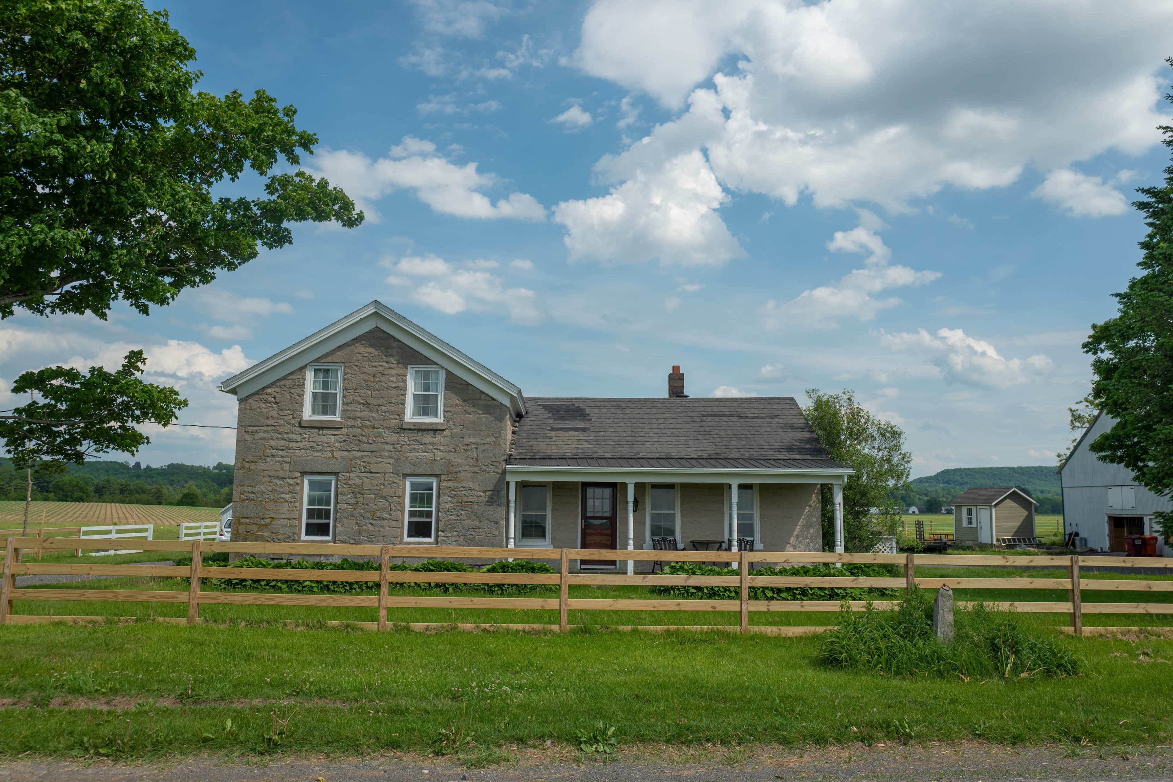 A two-story stone farmhouse with a porch is shown beside a wooden fence, set against a backdrop of green fields and cloudy skies.