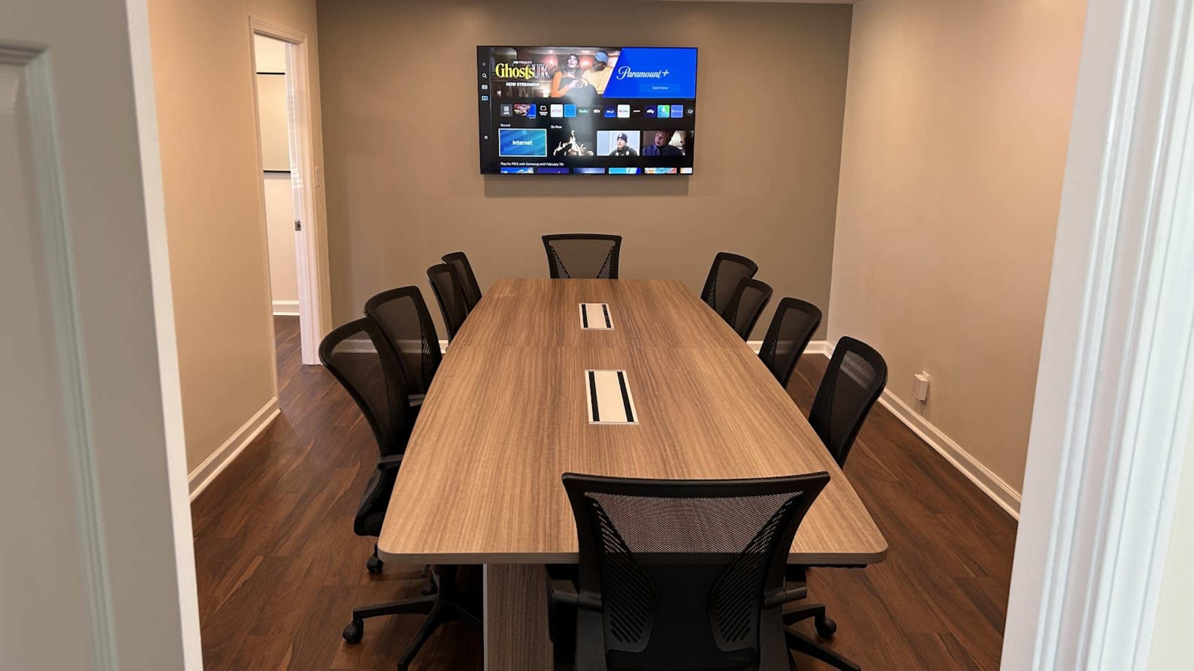A conference room features a large wooden table surrounded by eight black office chairs, with a wall-mounted screen displaying a video call interface.