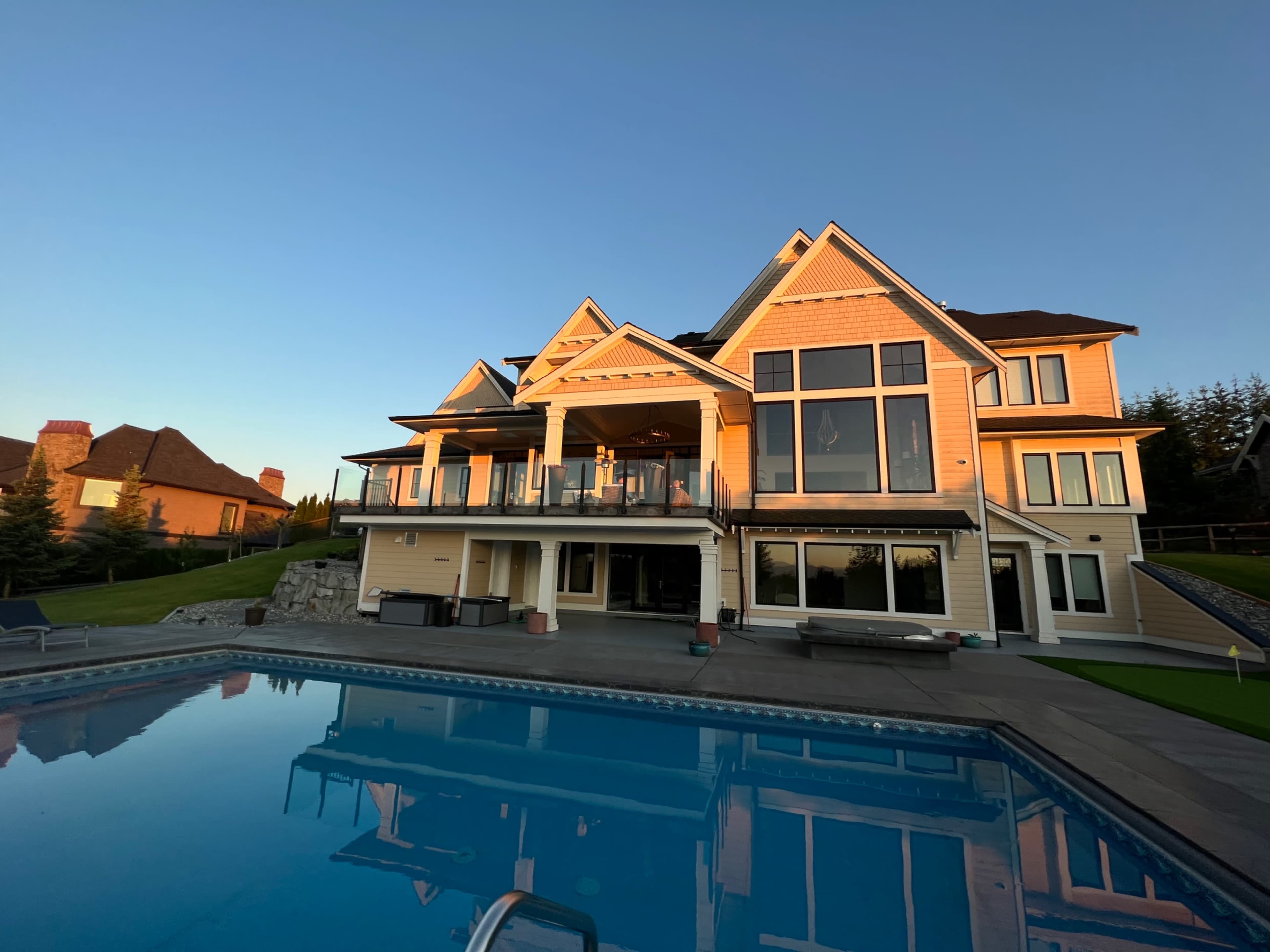 A large, modern house with multiple gables and a spacious deck overlooks a swimming pool, reflecting the clear sky.