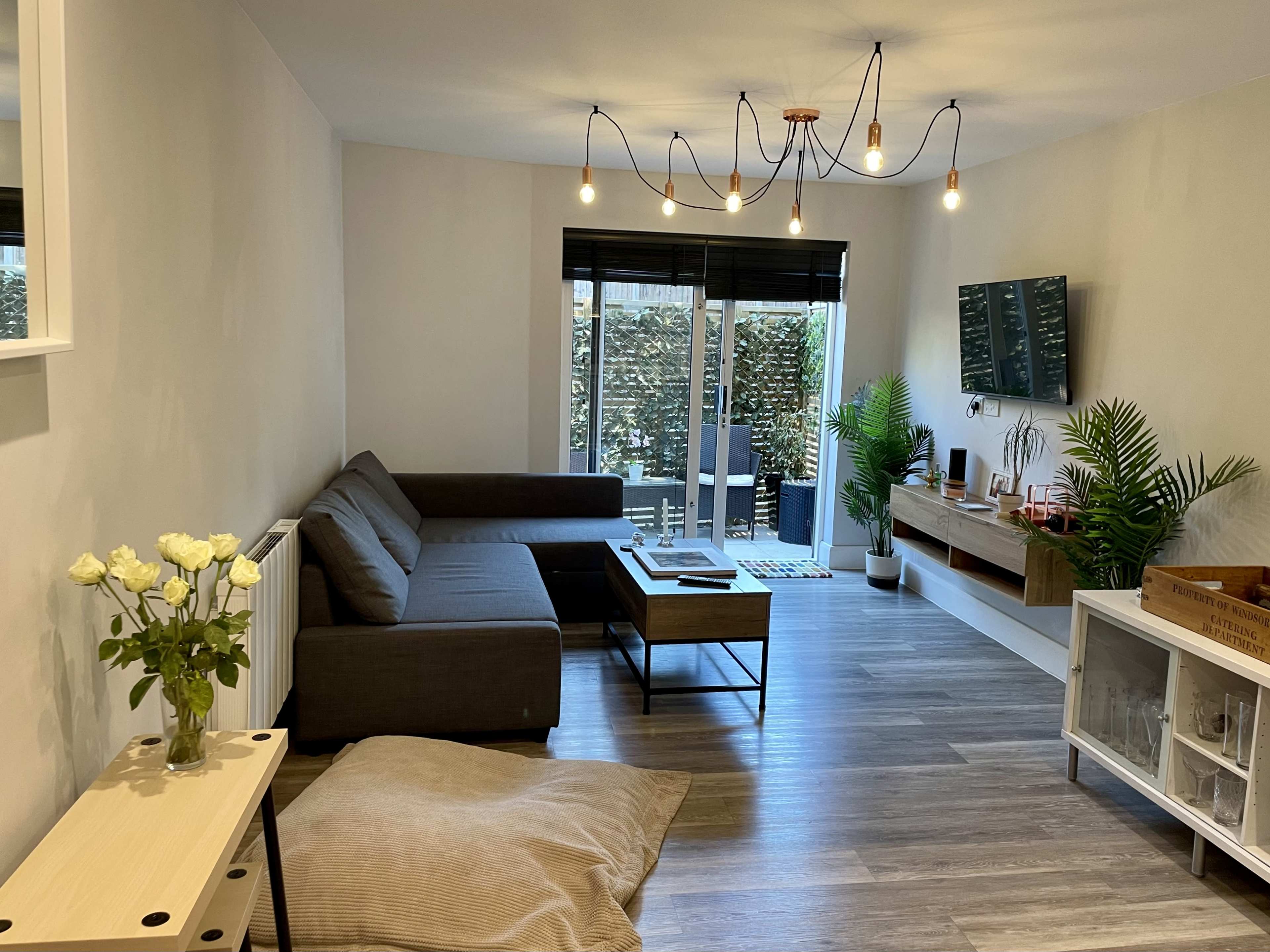 A modern living room features a gray sofa, a wooden coffee table, and a television mounted on the wall, with large windows allowing natural light and plants adding greenery to the space.