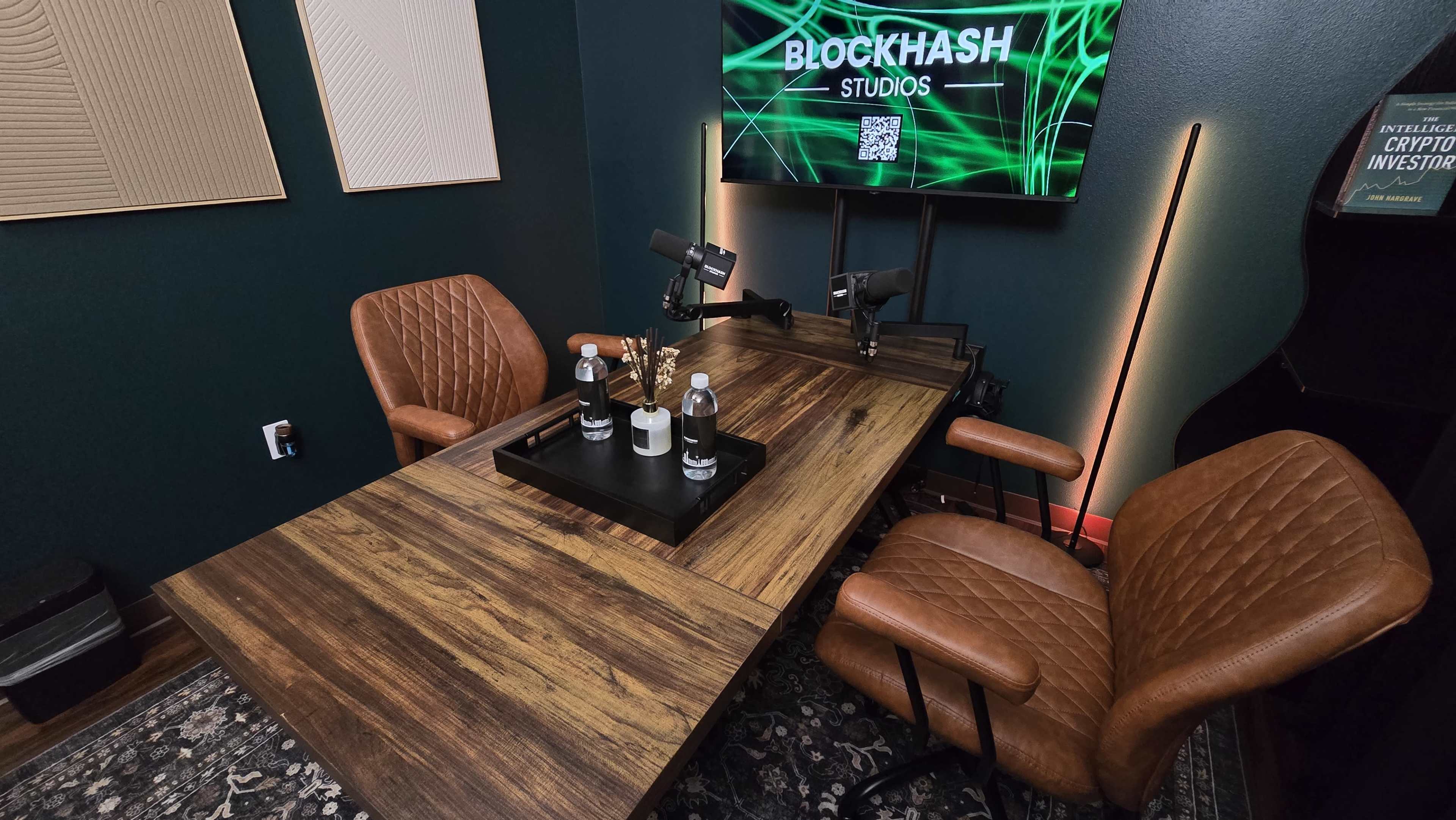 The image shows a modern studio setup with a wooden table, two leather chairs, and a television screen displaying "BLOCKHASH Studios."