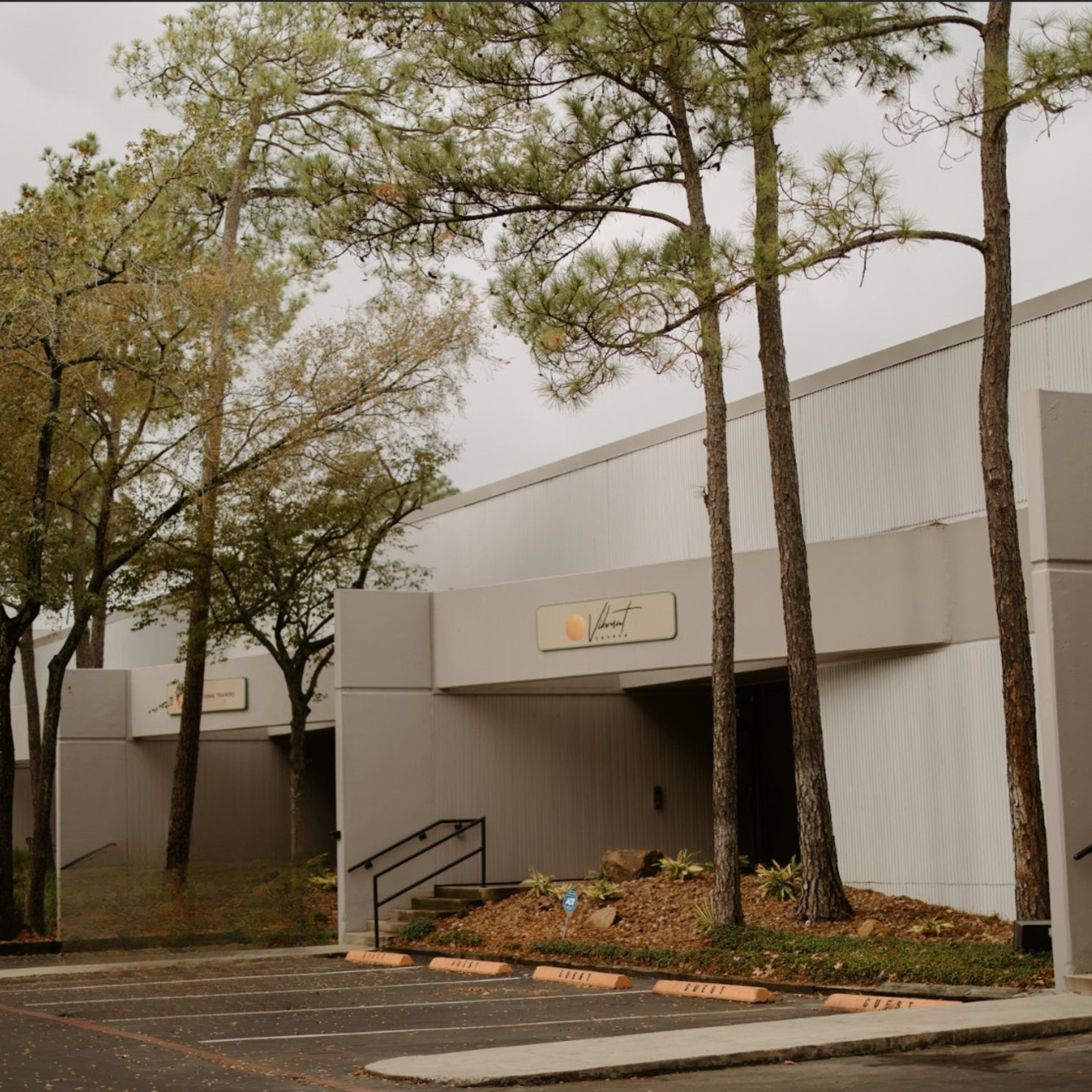 The image shows a gray commercial building with large windows and a sign, surrounded by tall pine trees and landscaped foliage.