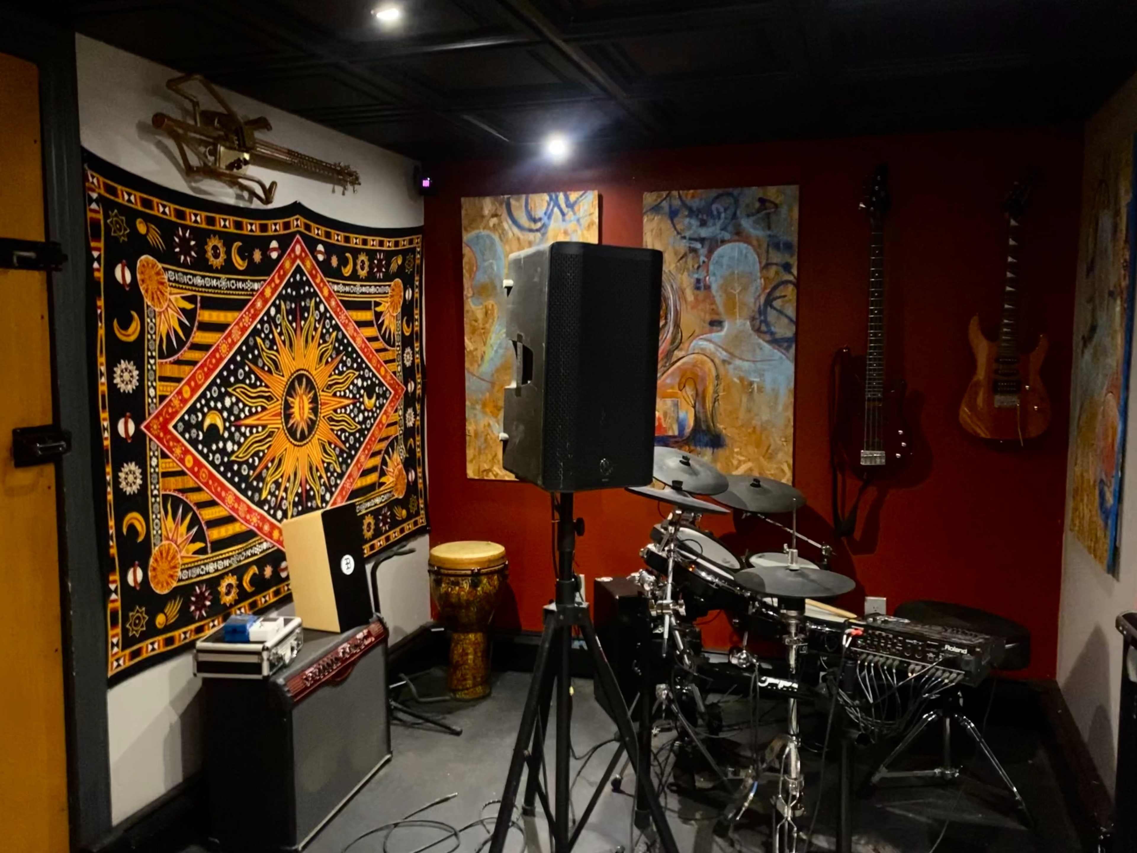 A music practice room featuring a drum set, an amplifier, a tapestry on the wall, and various musical instruments hanging on the red-painted walls.