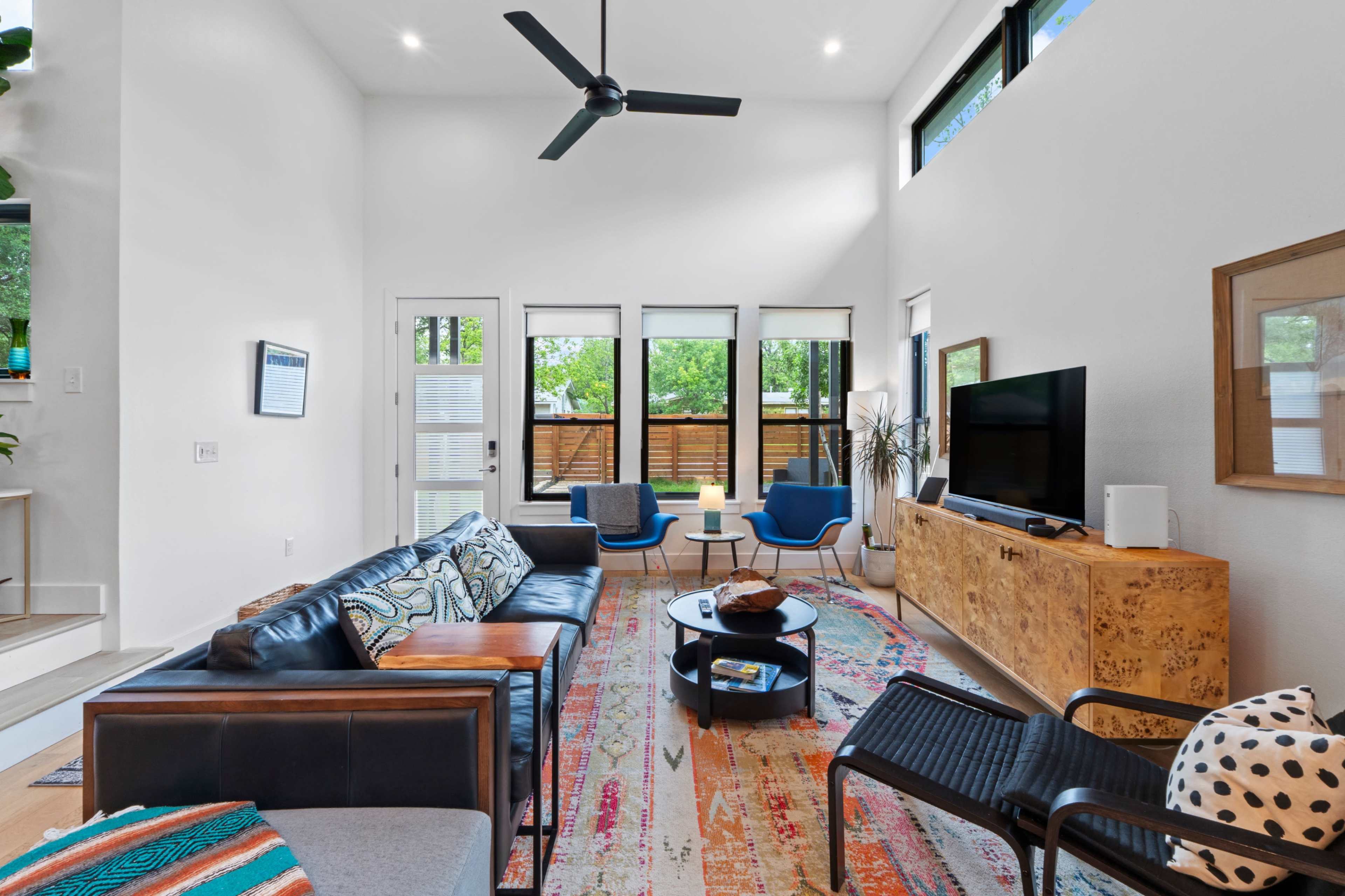 The living room features a modern design with a black leather sofa, two blue chairs, a wooden media console, and a colorful area rug.