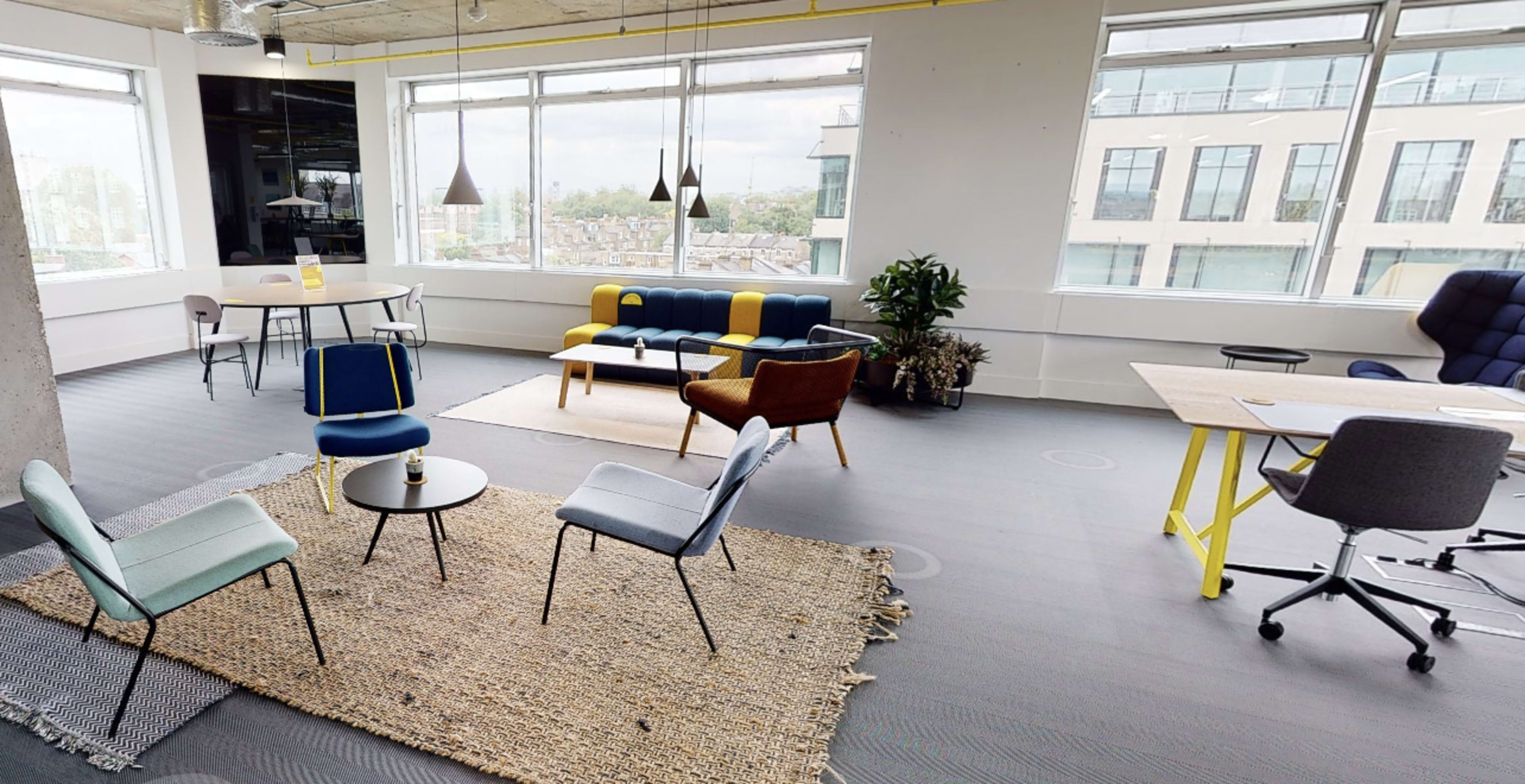 A modern office interior featuring a mix of seating areas, a small table, a desk, and large windows providing natural light.