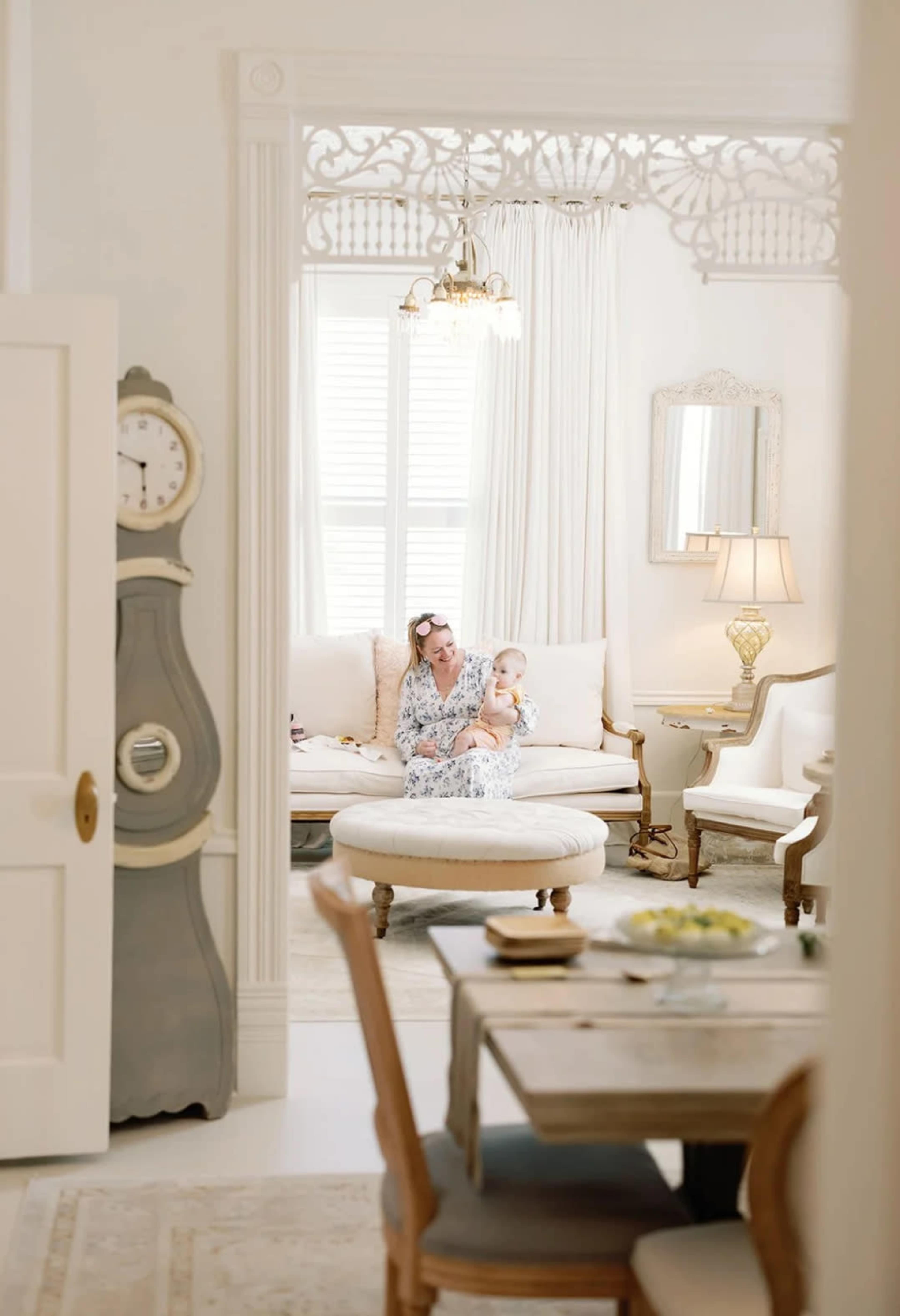 A woman seated on a round ottoman holds a baby in a bright, elegantly decorated room with light-colored furniture and large windows.
