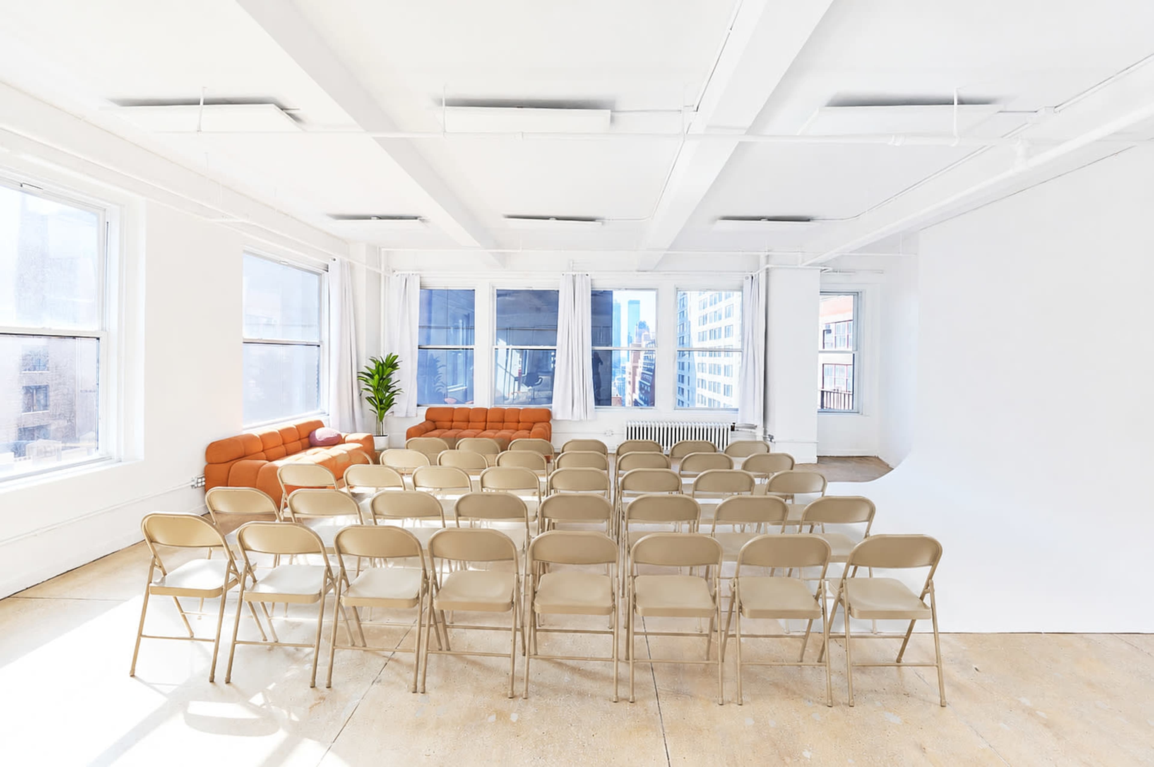 The image shows a brightly lit, spacious room with a row of beige folding chairs facing a white wall, and a small orange sofa positioned to the left near the windows.