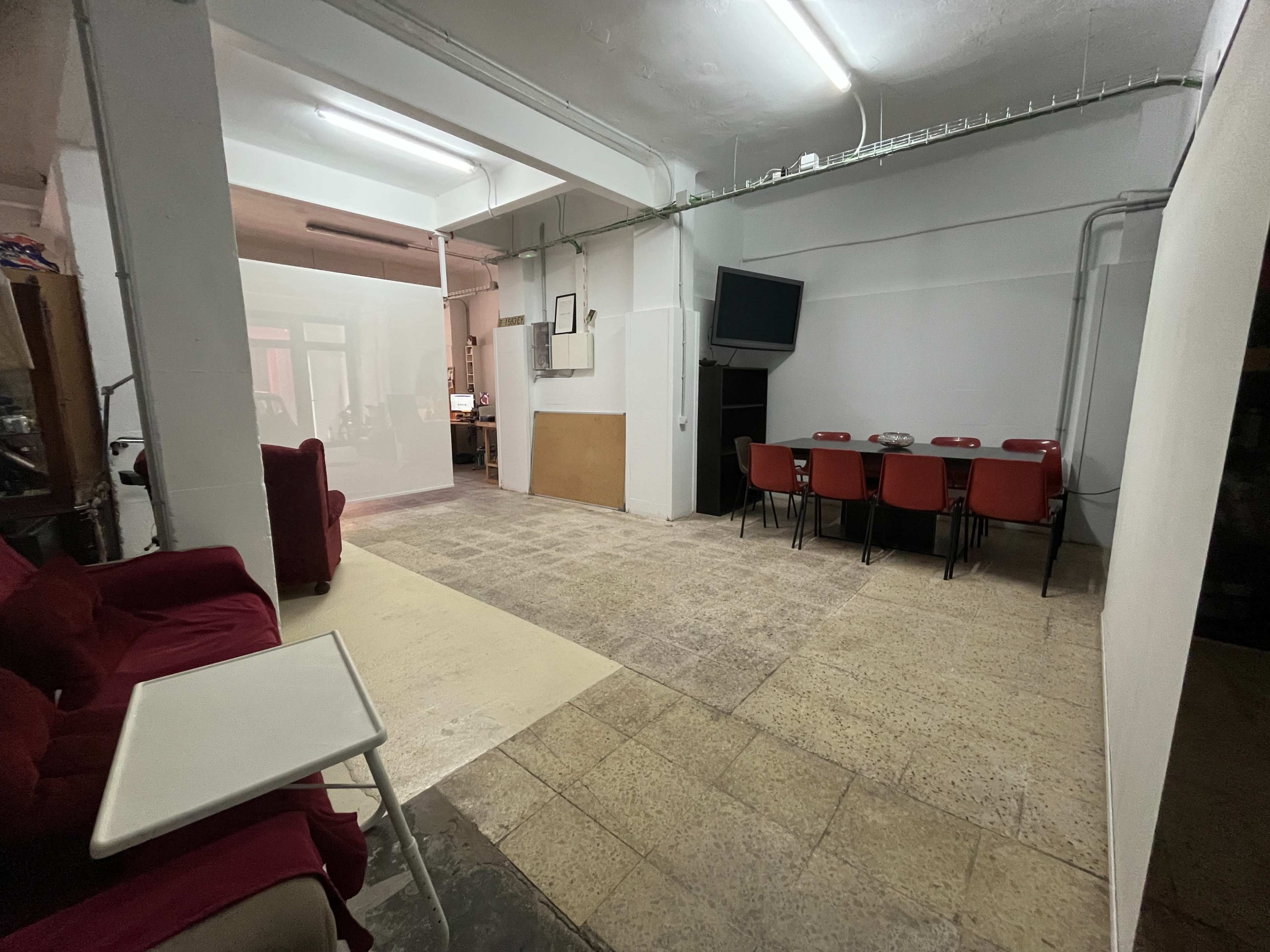 The image shows a spacious room with a seating area on the left and a long table surrounded by red chairs on the right, along with an adjoining area that appears to have computer workstations.