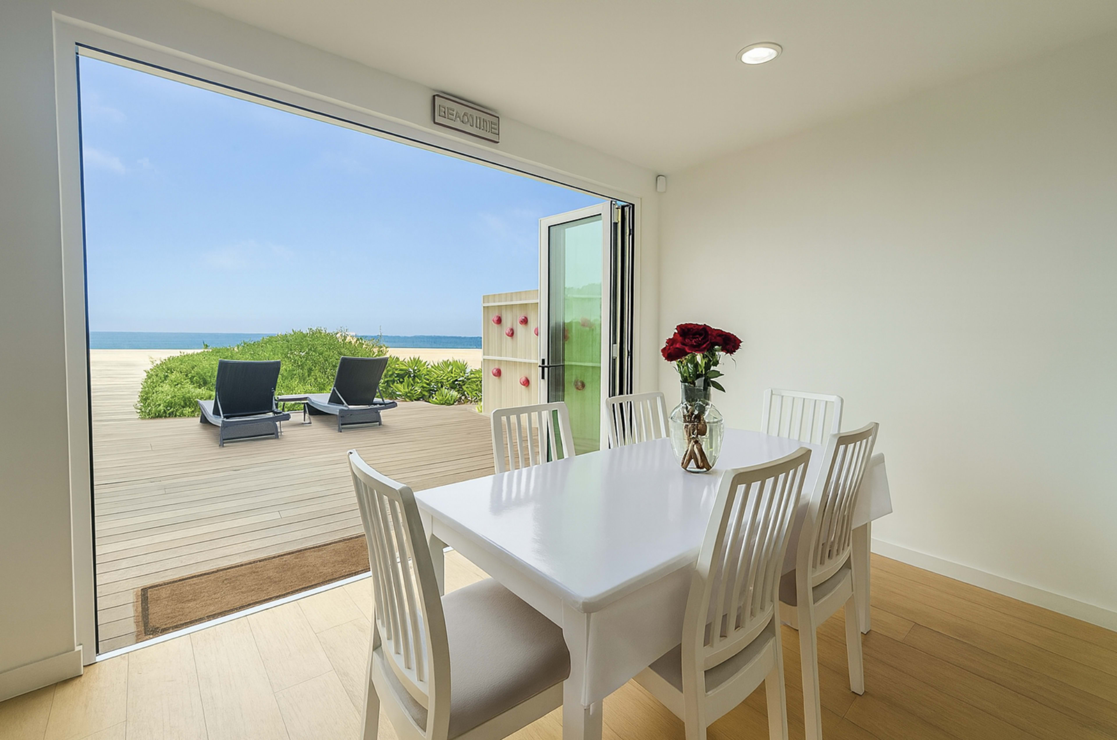 A dining area features a white table and chairs, with a vase of roses on the table, overlooking a patio and beach view through an open glass door.