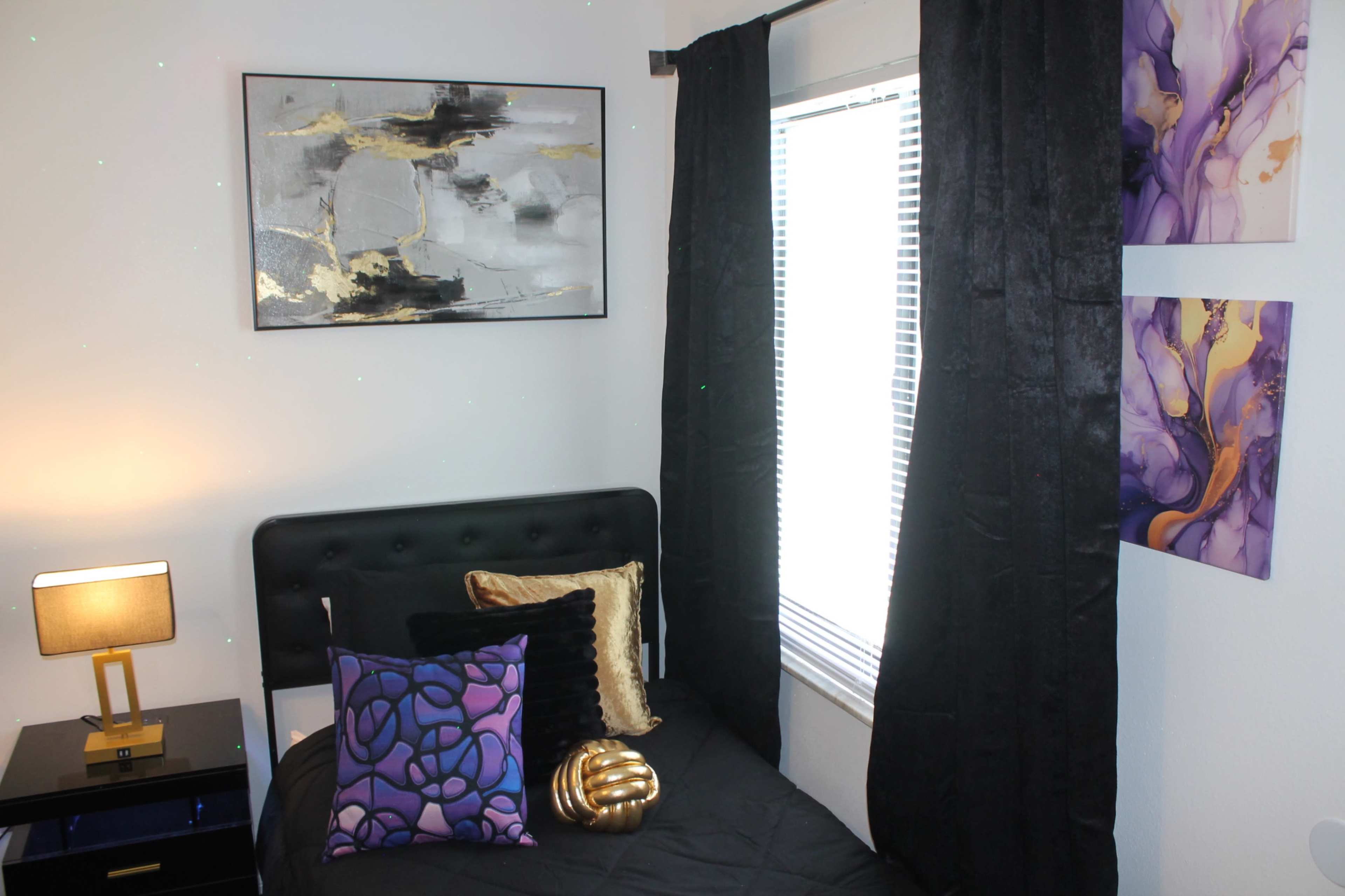 A small bedroom featuring a black bed with decorative pillows, a nightstand with a lamp, and artwork on the walls.