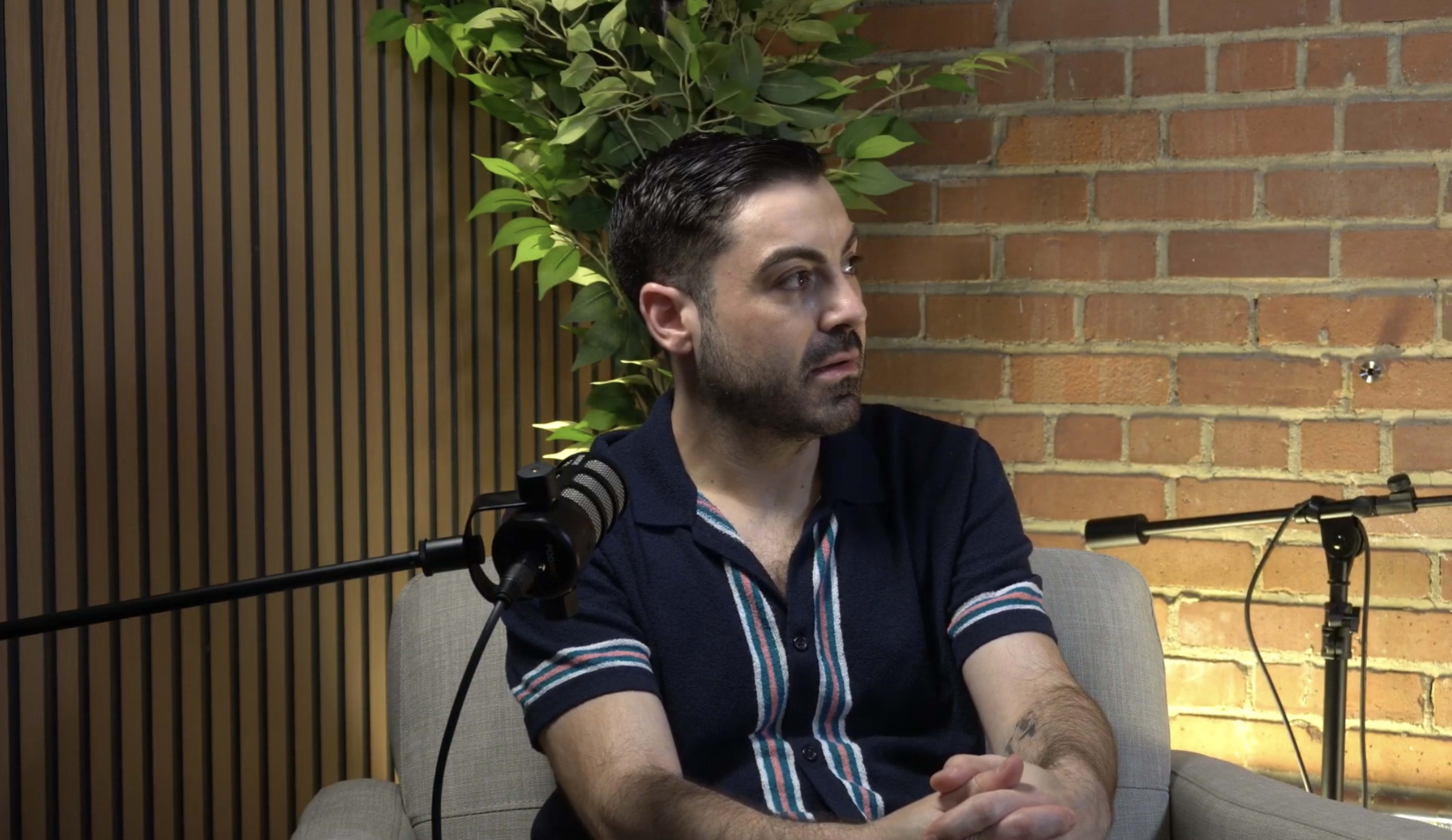 A man sits on a couch in a room with a brick wall and a plant in the background, while a microphone is positioned nearby.