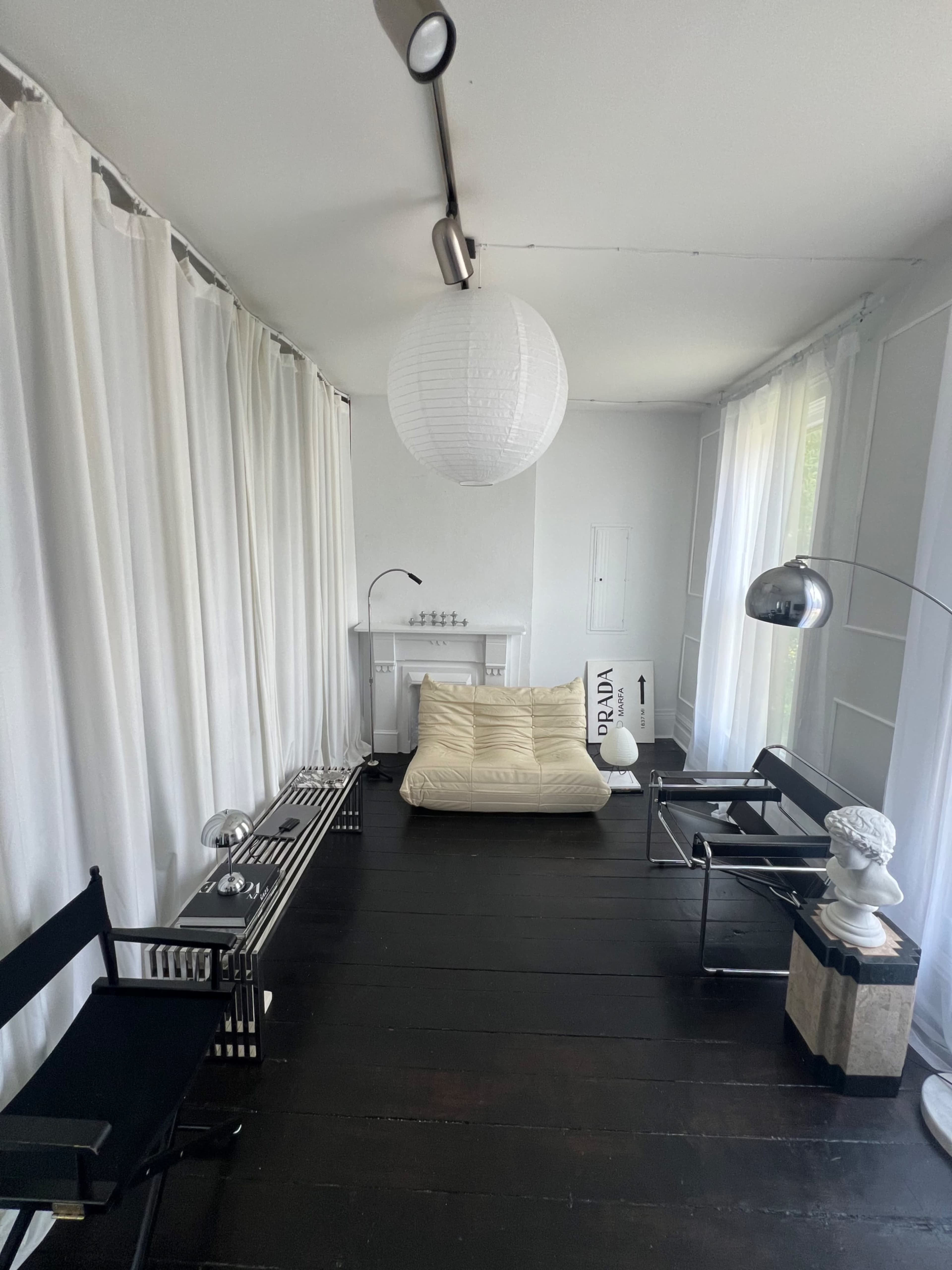 The image shows a minimalist room with white curtains, a large round light fixture, a beige floor cushion, and modern furniture including two chairs and a decorative statue.