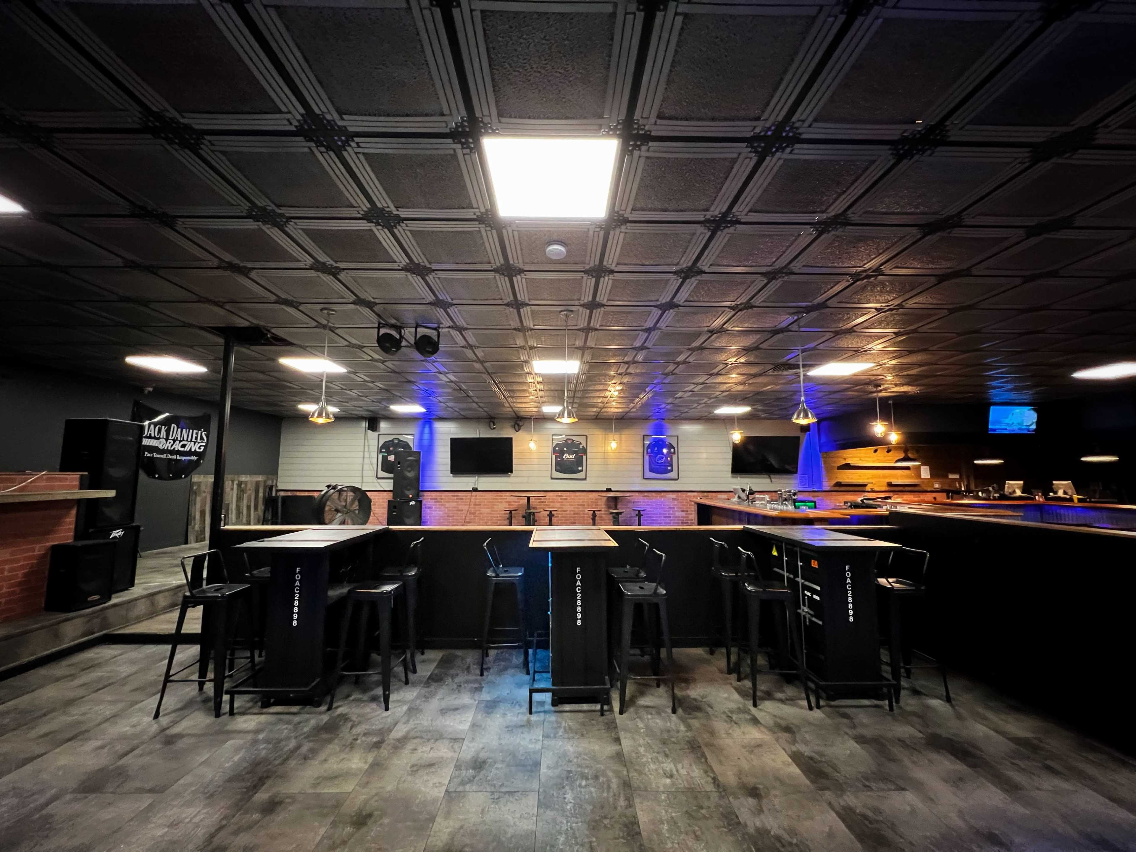 A modern bar area with a black ceiling, several high tables with stools, and illuminated features in a dimly lit space.