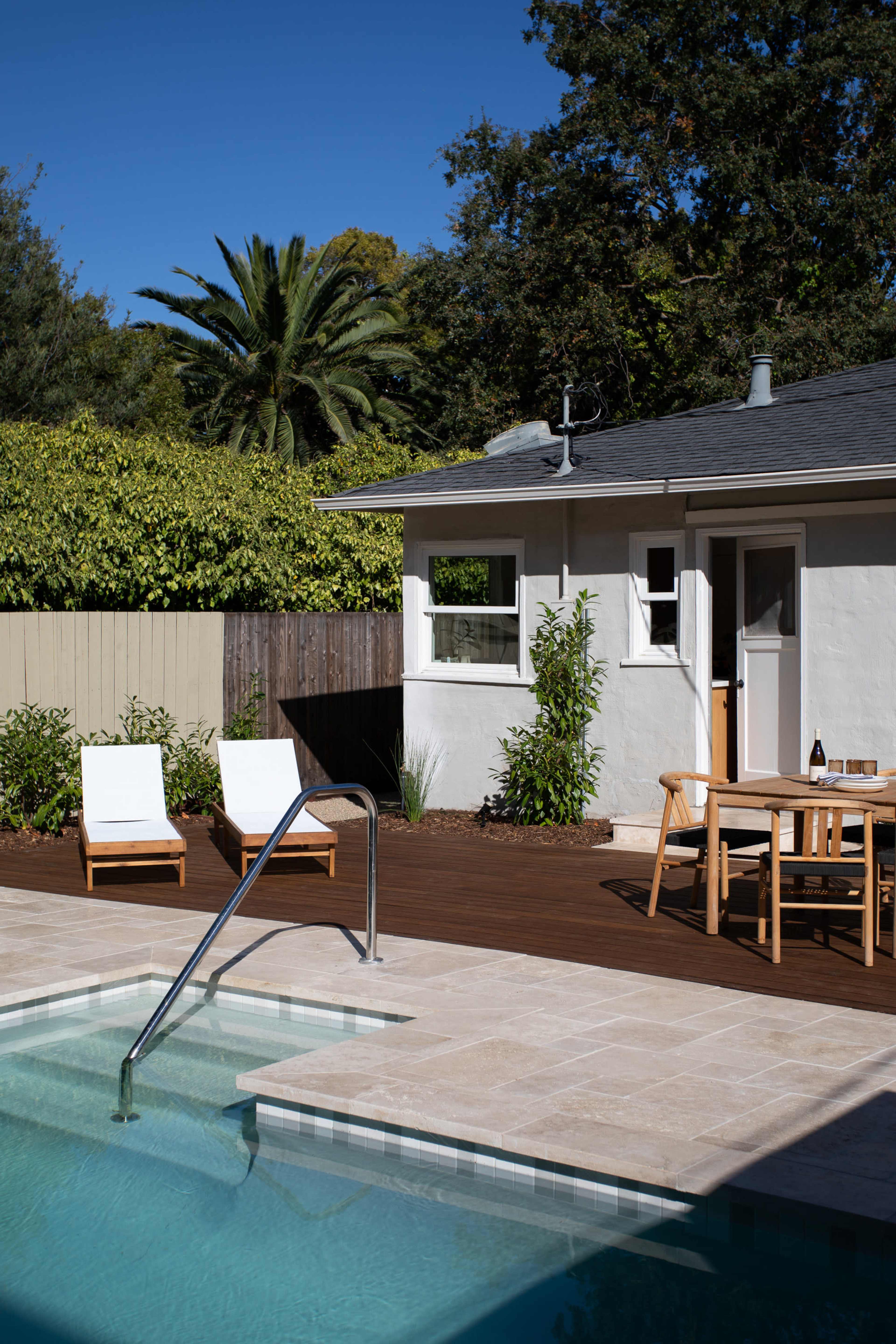 A clear swimming pool is surrounded by a wooden deck with two lounge chairs and a dining table set near a house and greenery.