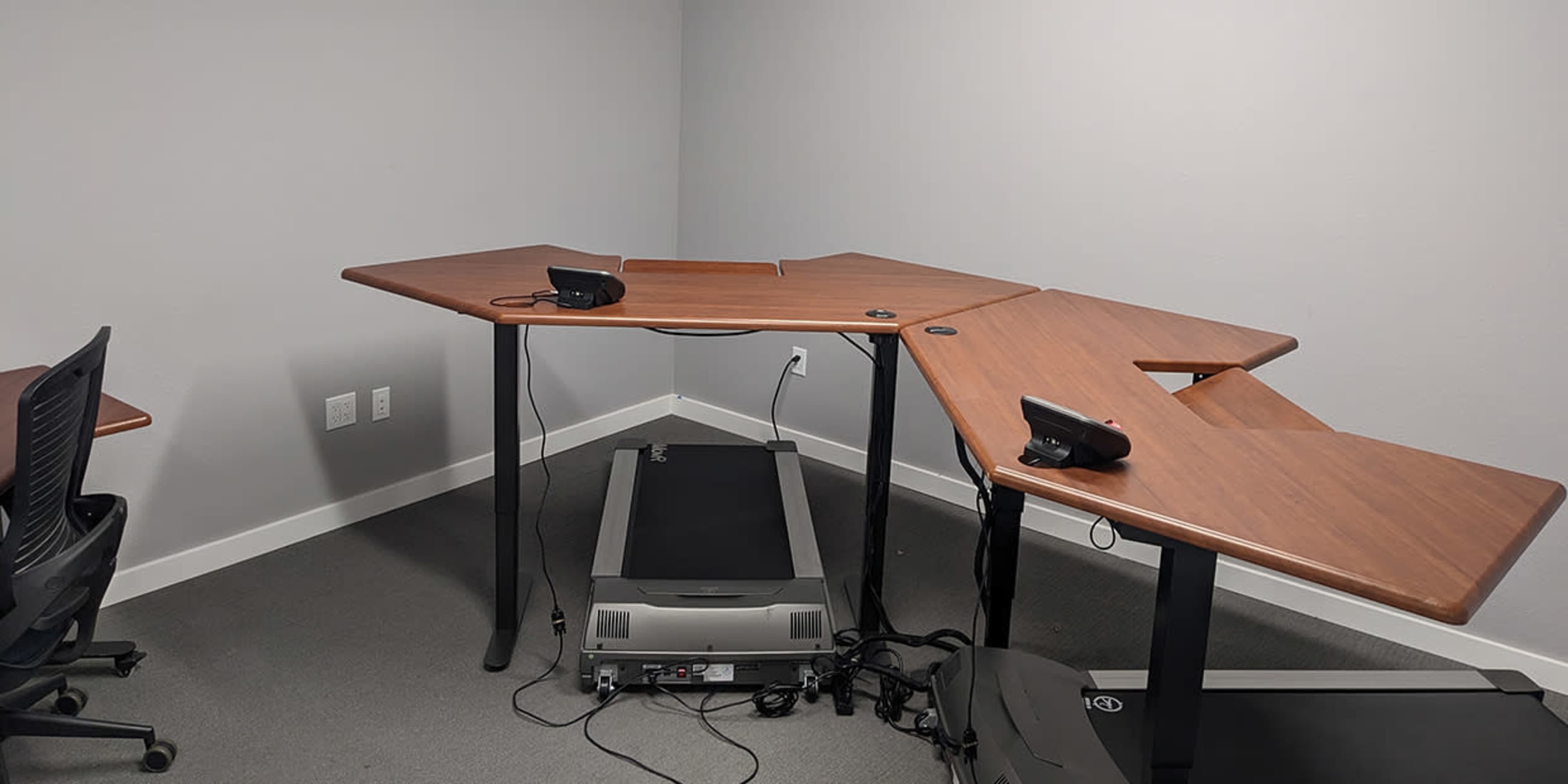 A multi-segmented wooden desk with telephone equipment sits in a corner of a room, next to a treadmill and black office chairs.