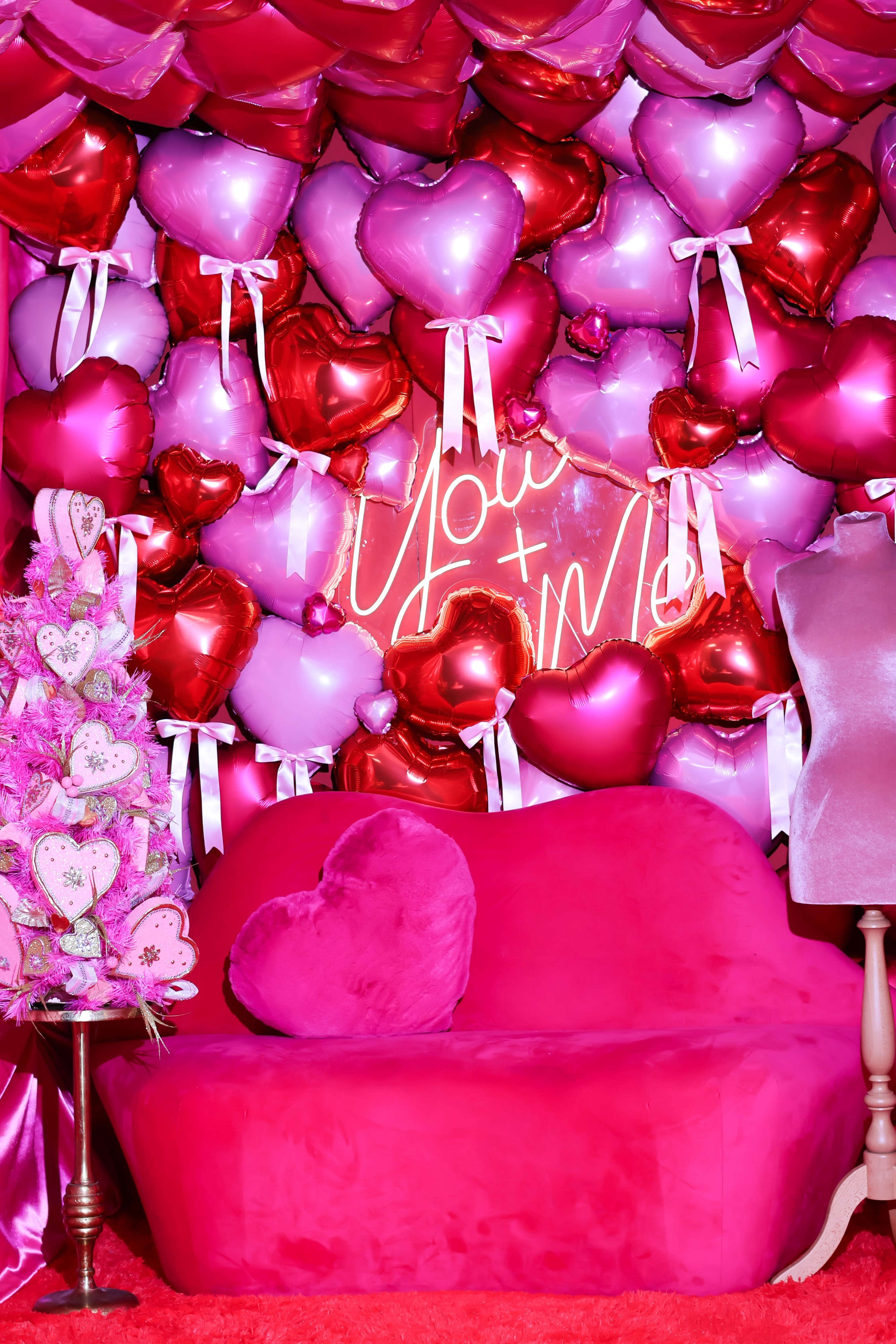 The image shows a vibrant, pink-themed room decorated with heart-shaped balloons and a neon sign that says "You + Me," along with a plush pink couch and a decorative tree.