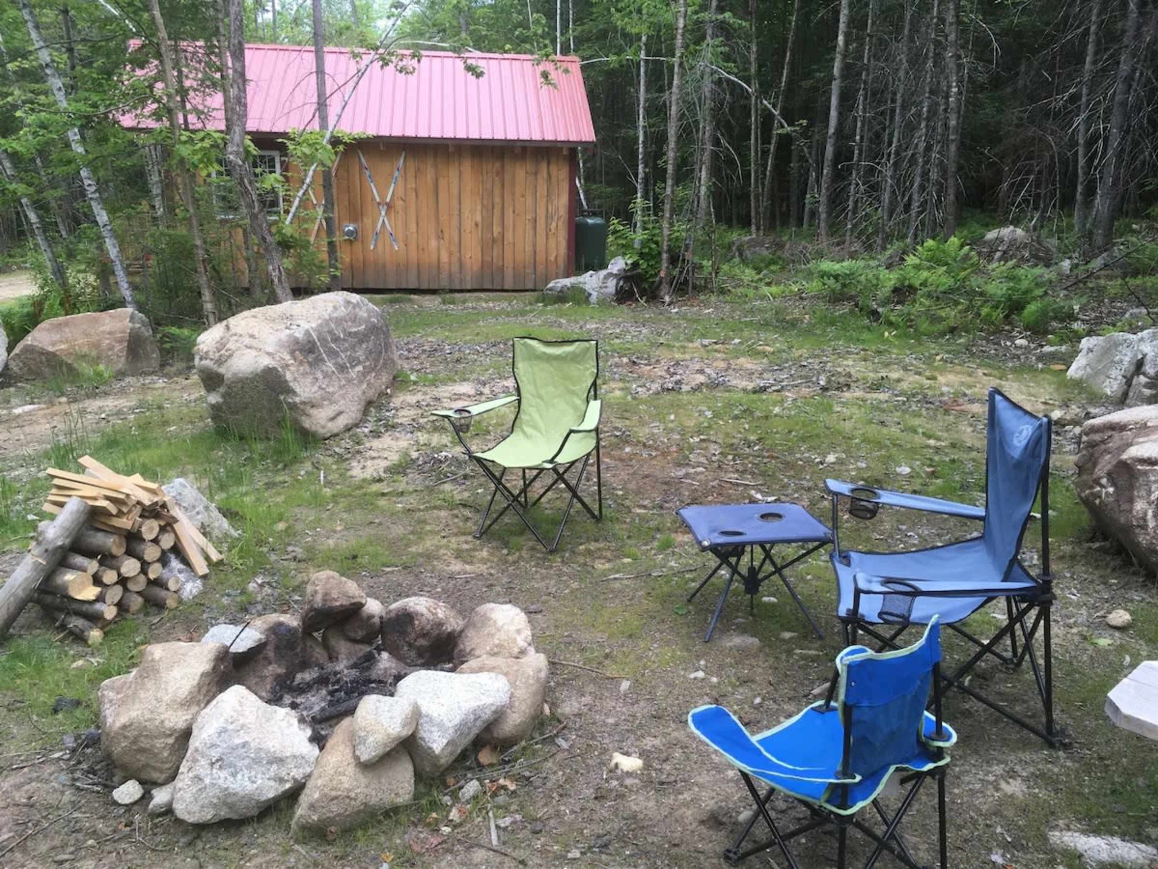 A campfire area with several folding chairs set around a stone fire pit, next to a wooden shed in a wooded environment.