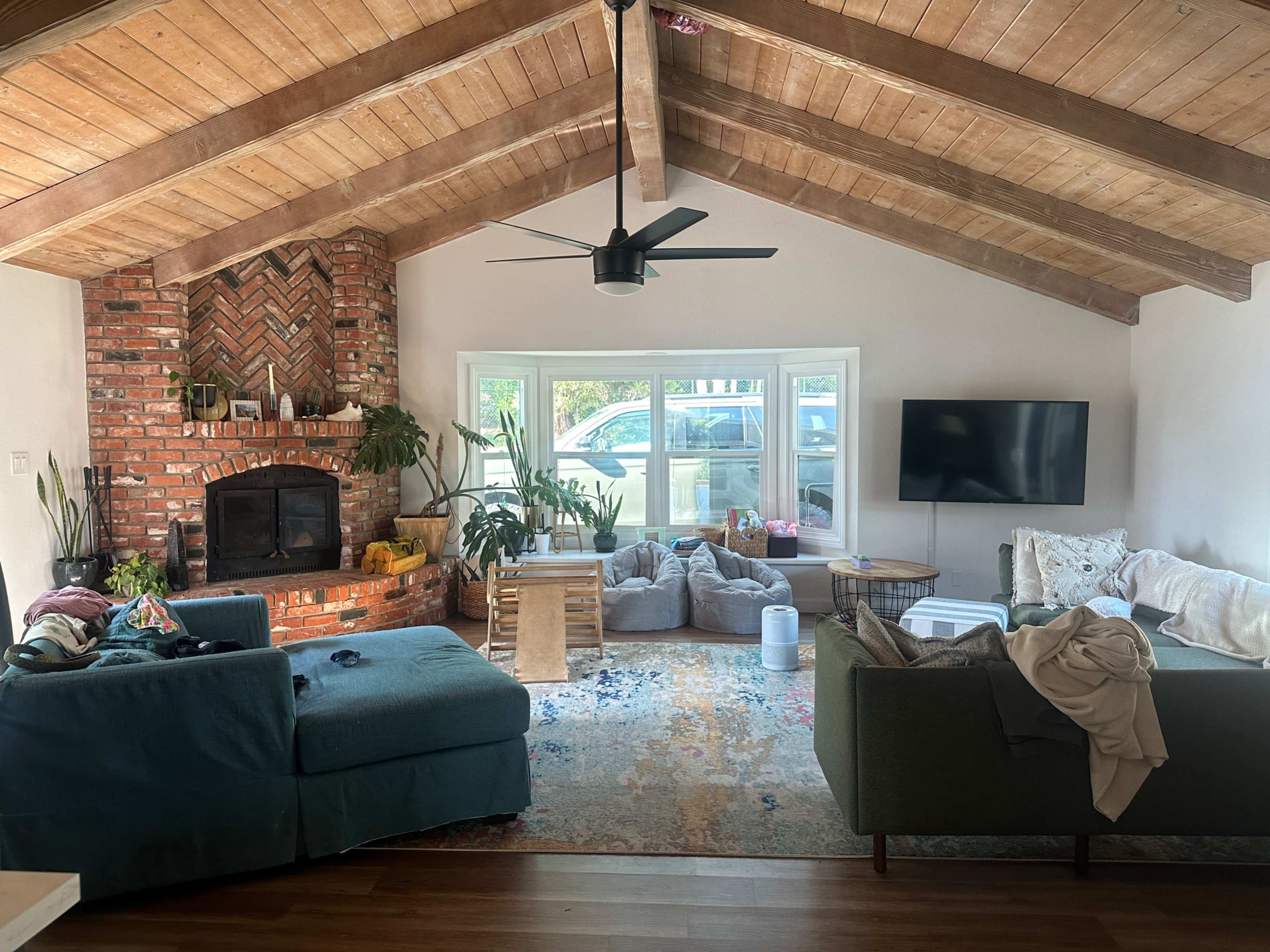 The living room features a high, exposed beam ceiling, a brick fireplace, large windows with outdoor views, and a mix of comfortable seating arranged around a colorful area rug.