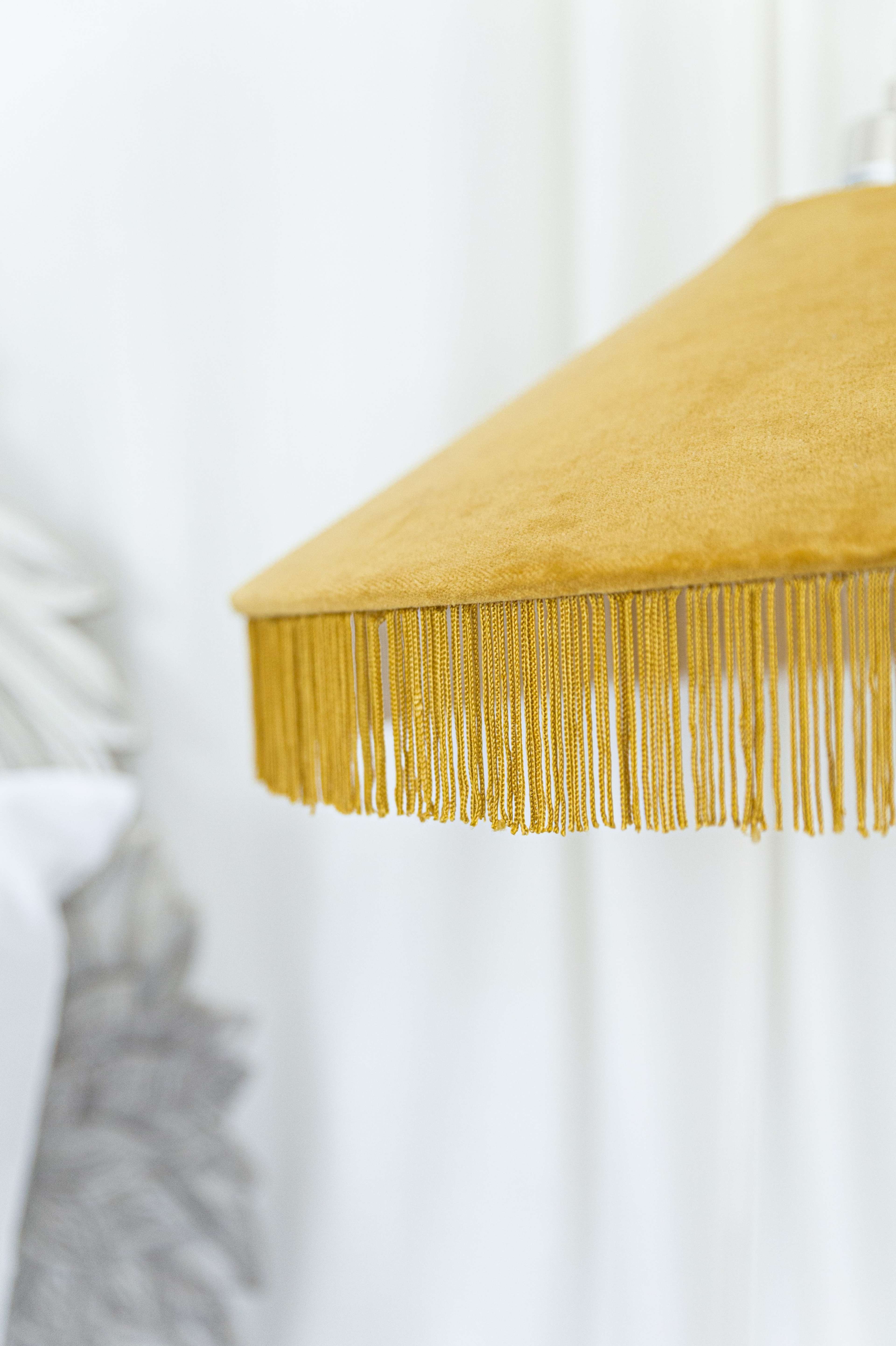A mustard-colored lamp shade with fringe hangs above a soft, textured background.
