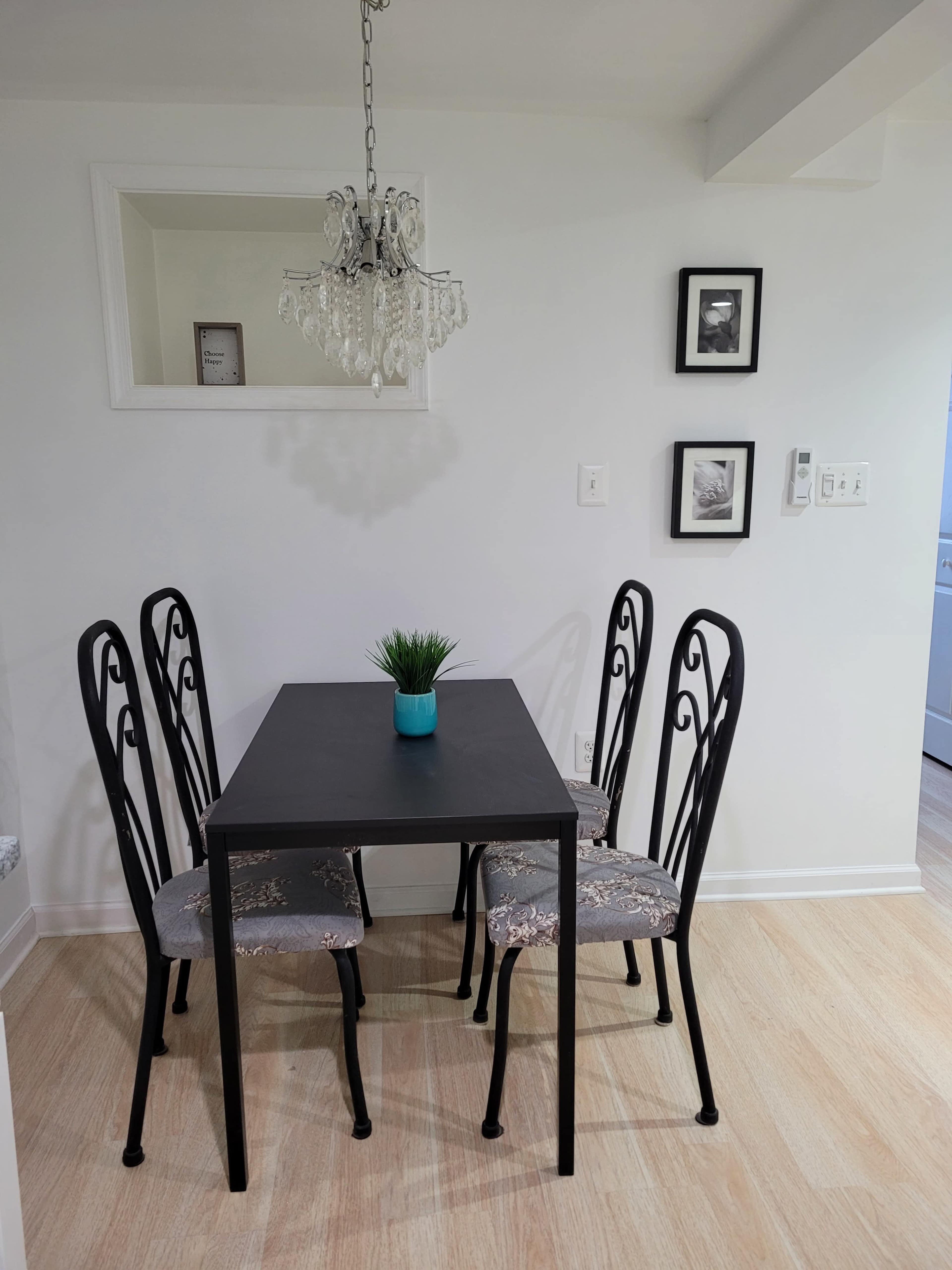 A small dining area features a black table surrounded by four wrought iron chairs with patterned cushions, complemented by a potted plant on the table and a chandelier overhead.