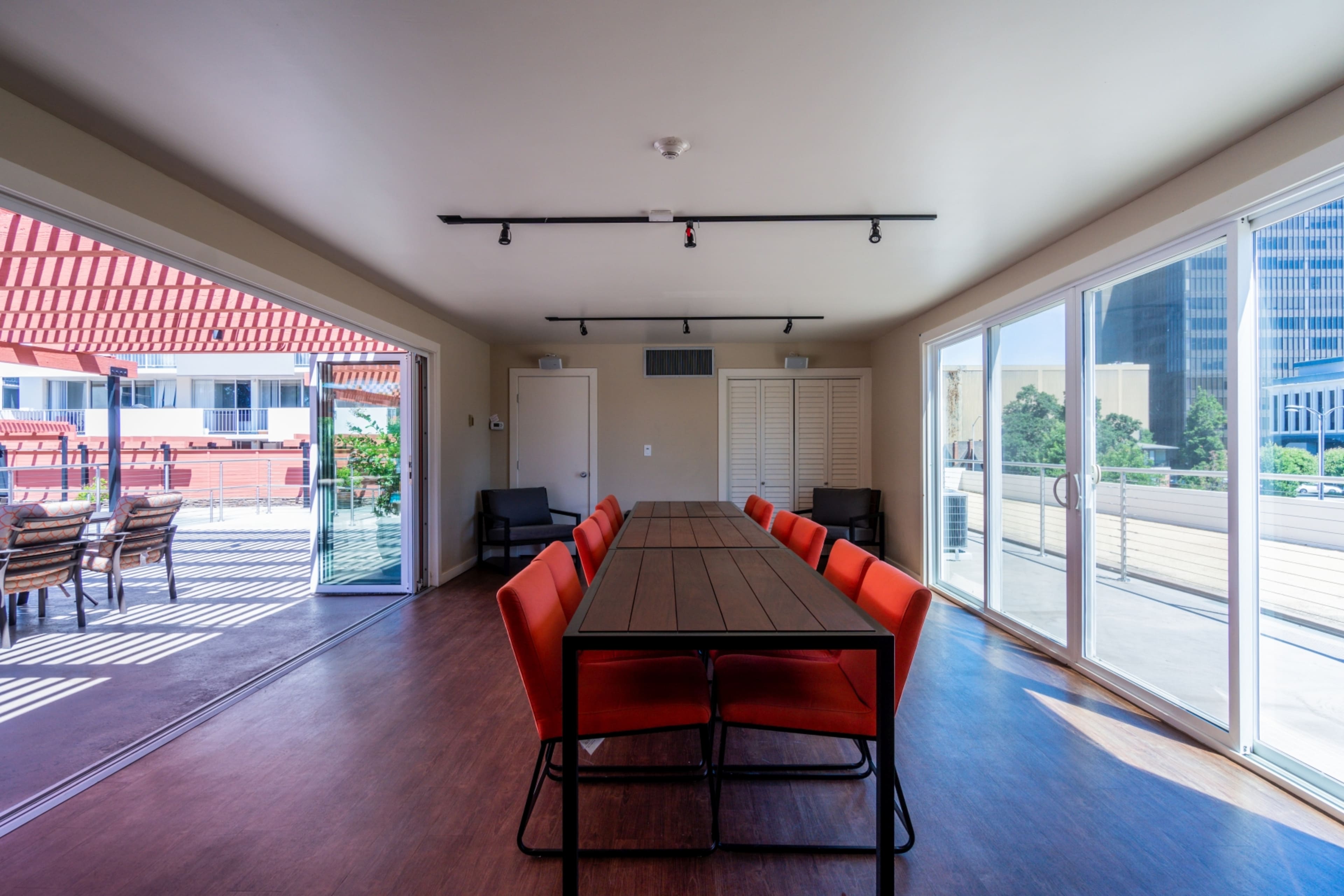 A long wooden table with orange chairs is set up in a brightly lit room with large sliding glass doors leading to an outdoor area.
