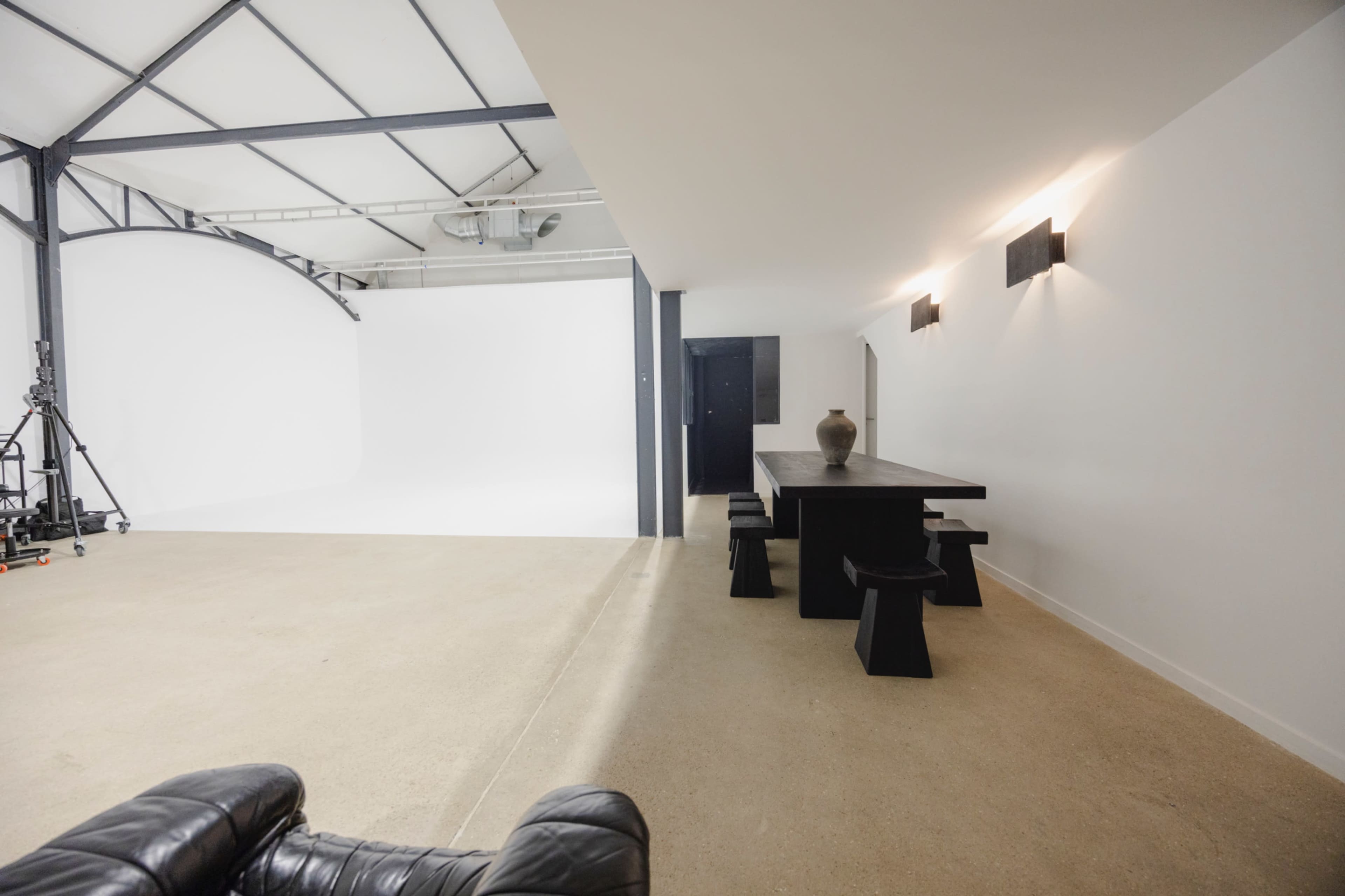 The image shows a spacious, minimalist studio featuring a long dark dining table with stools, a white backdrop for photography, and industrial-style lighting.