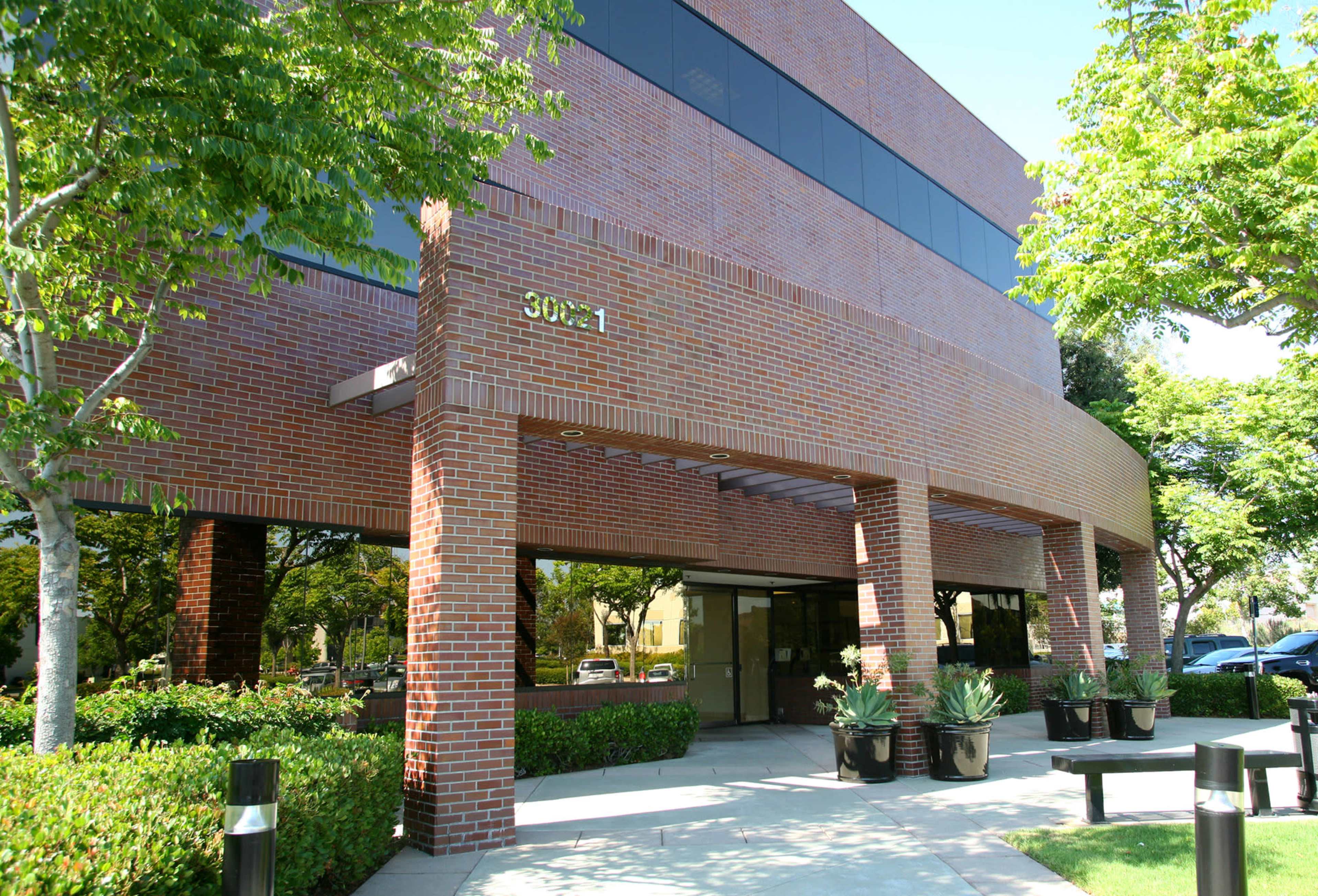 The image shows a modern brick office building surrounded by trees and landscaped greenery.