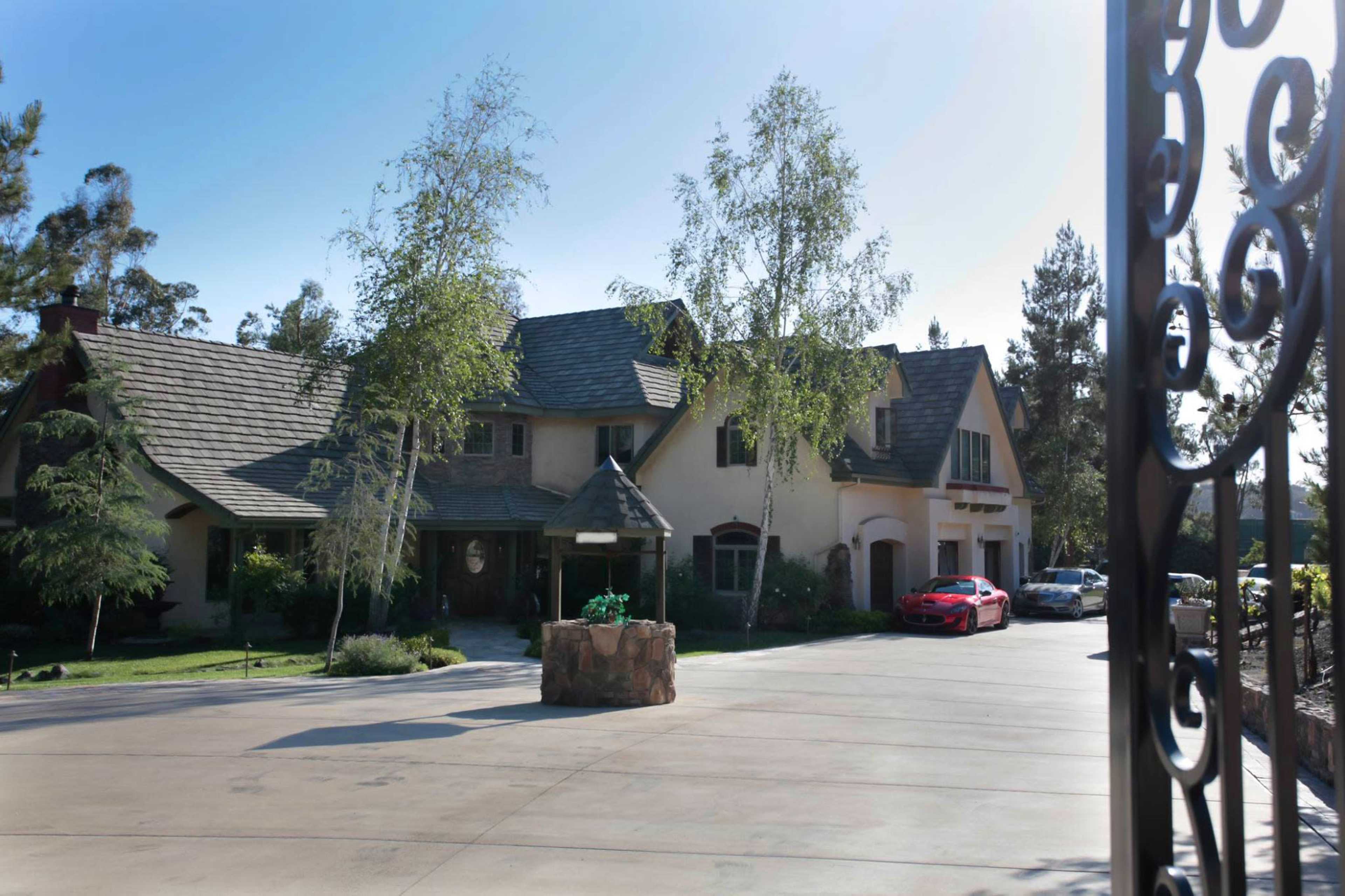 A large house with a gray roof and beige exterior is set back from a concrete driveway, surrounded by trees and greenery.