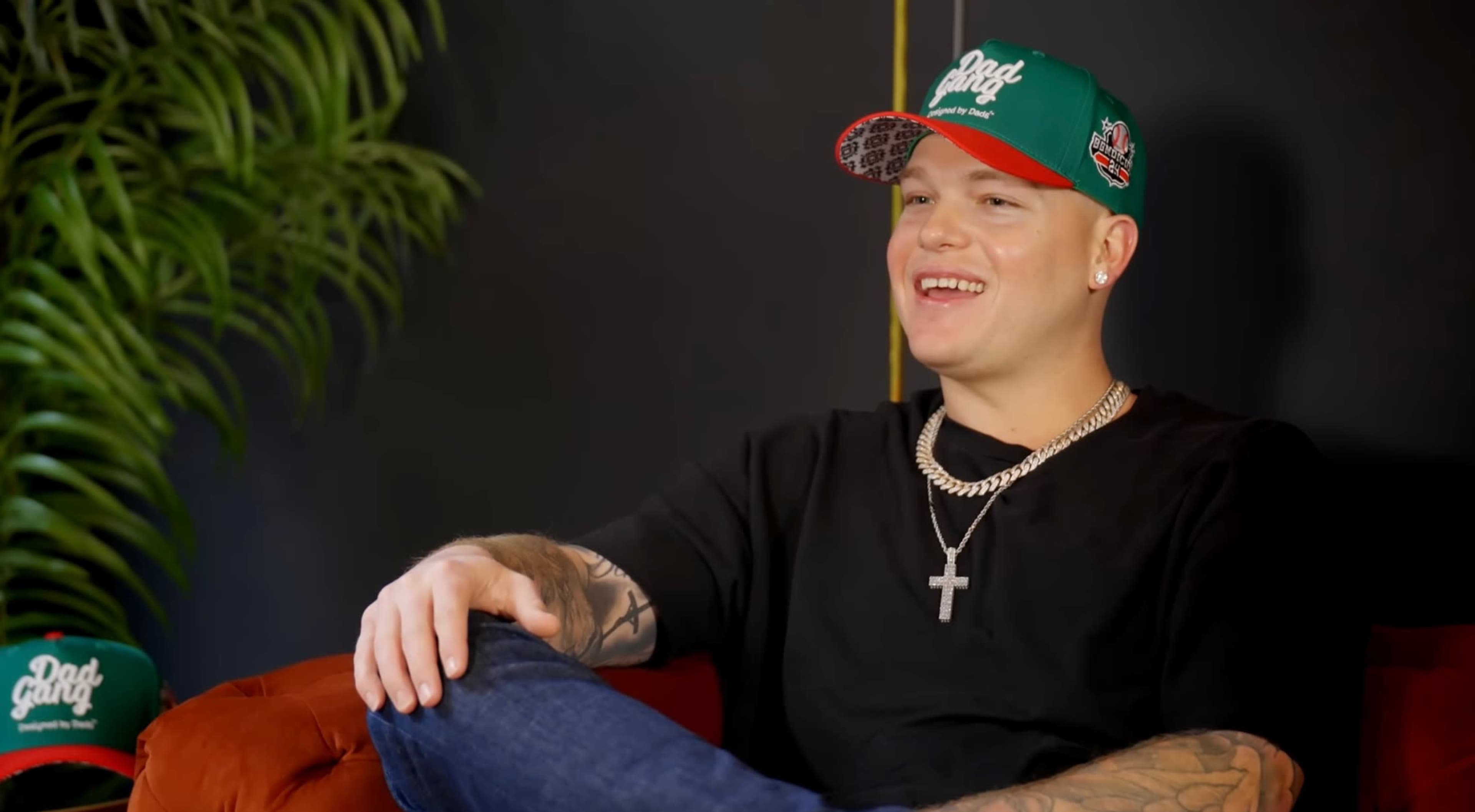 A man wearing a green and red cap sits comfortably on a couch, displaying tattoos on his arms and a cross necklace.